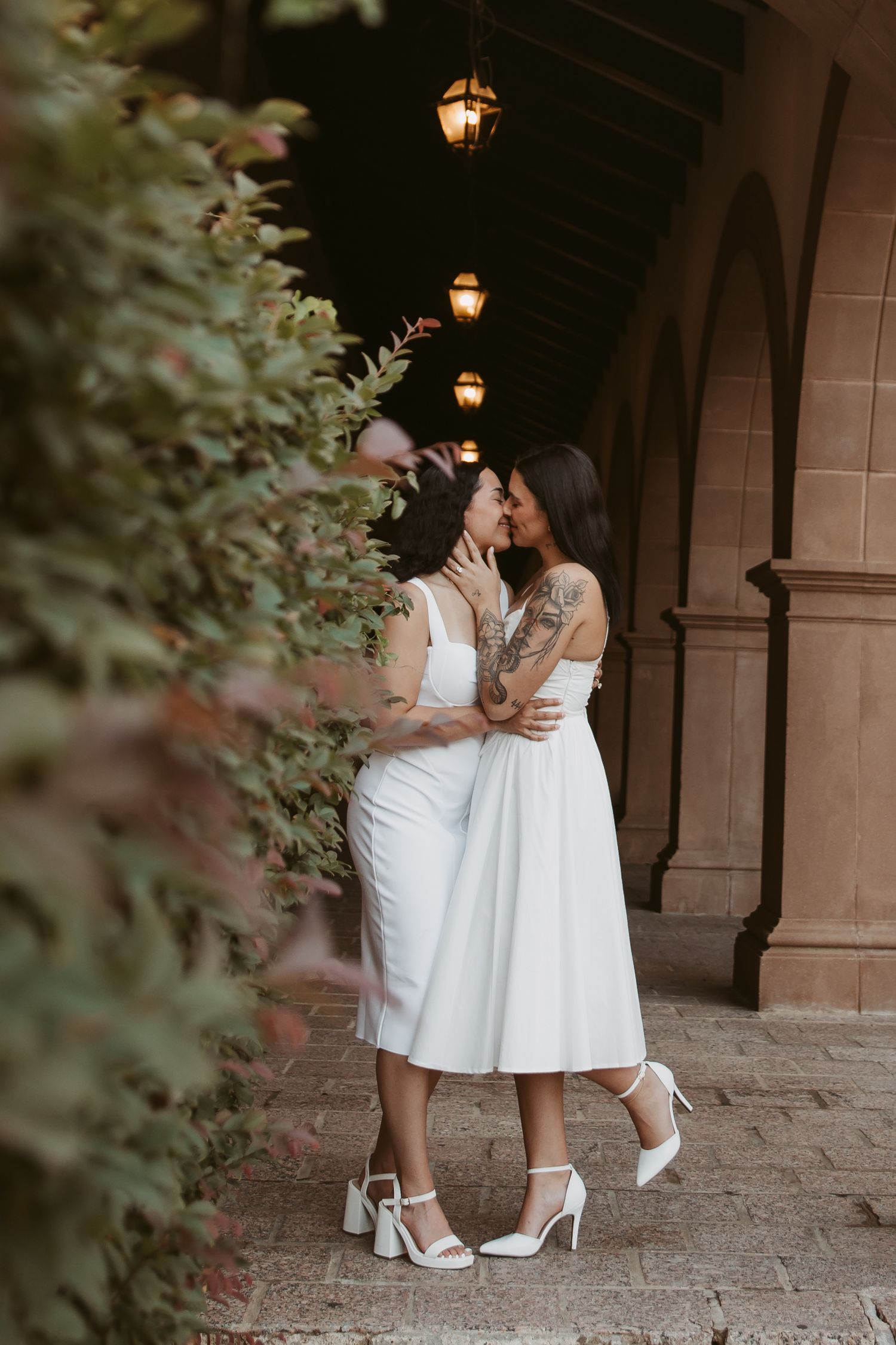 Two brides in white dresses embrace and kiss under arched walkway with warm pendant lights.