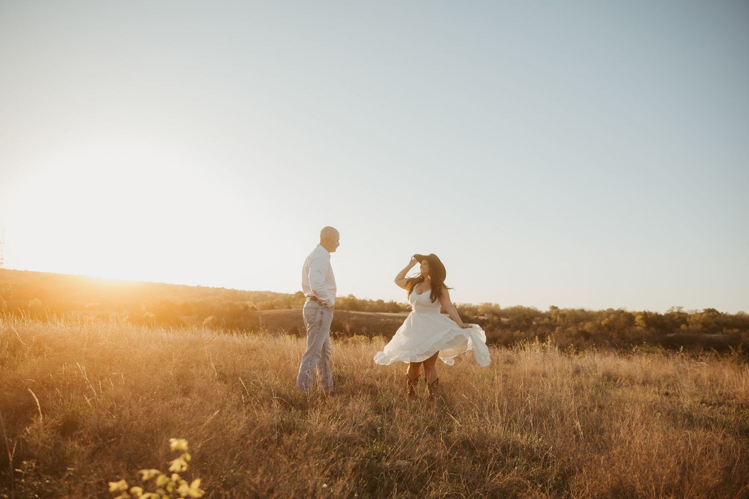Couple in white outfits dancing in golden sunlit field at sunset, romantic outdoor engagement photo.