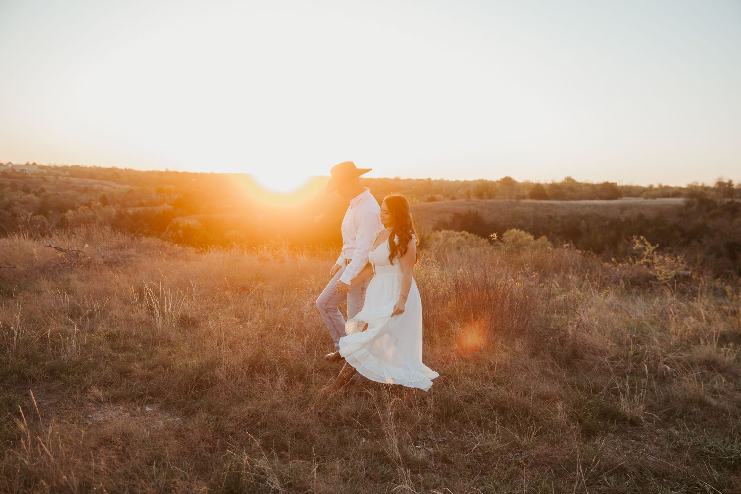 Couple dancing in golden sunset light in a field, woman in white dress and man in cowboy hat.