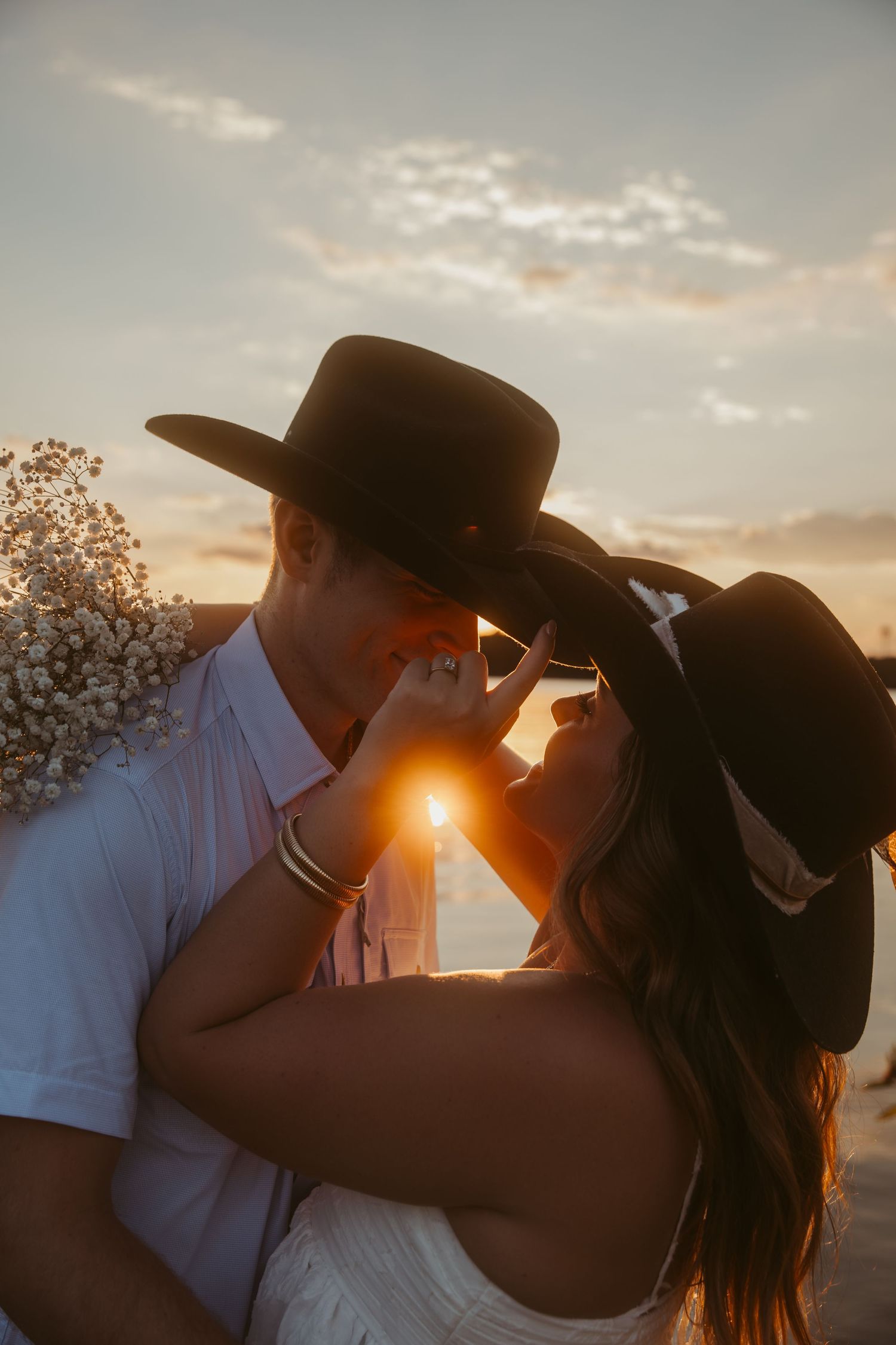Couple in cowboy hats sharing a romantic kiss at golden sunset, with warm backlight and floral bouquet.