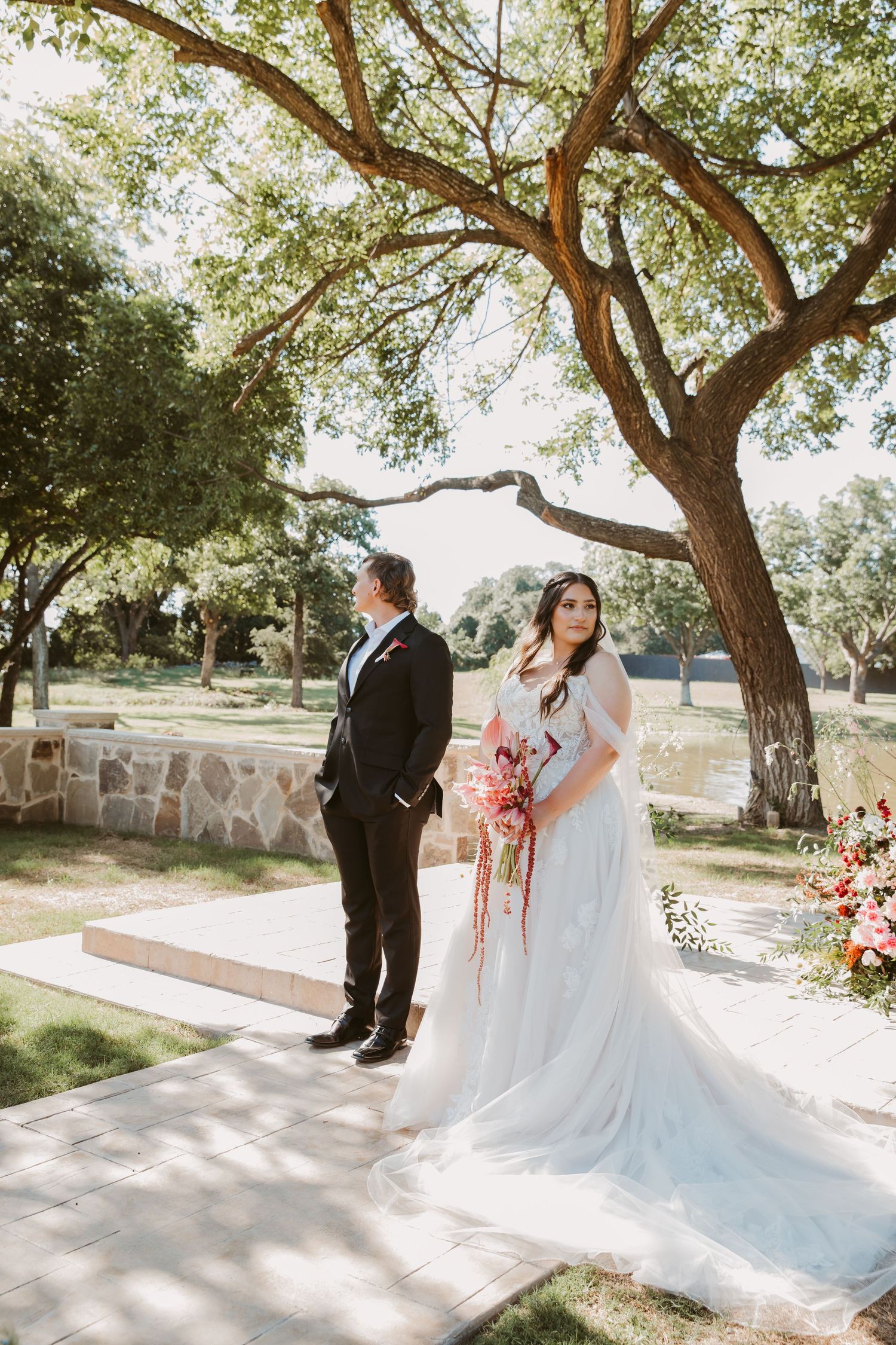 Bride in white gown holding bouquet stands with groom in black tux under oak trees at outdoor wedding venue.