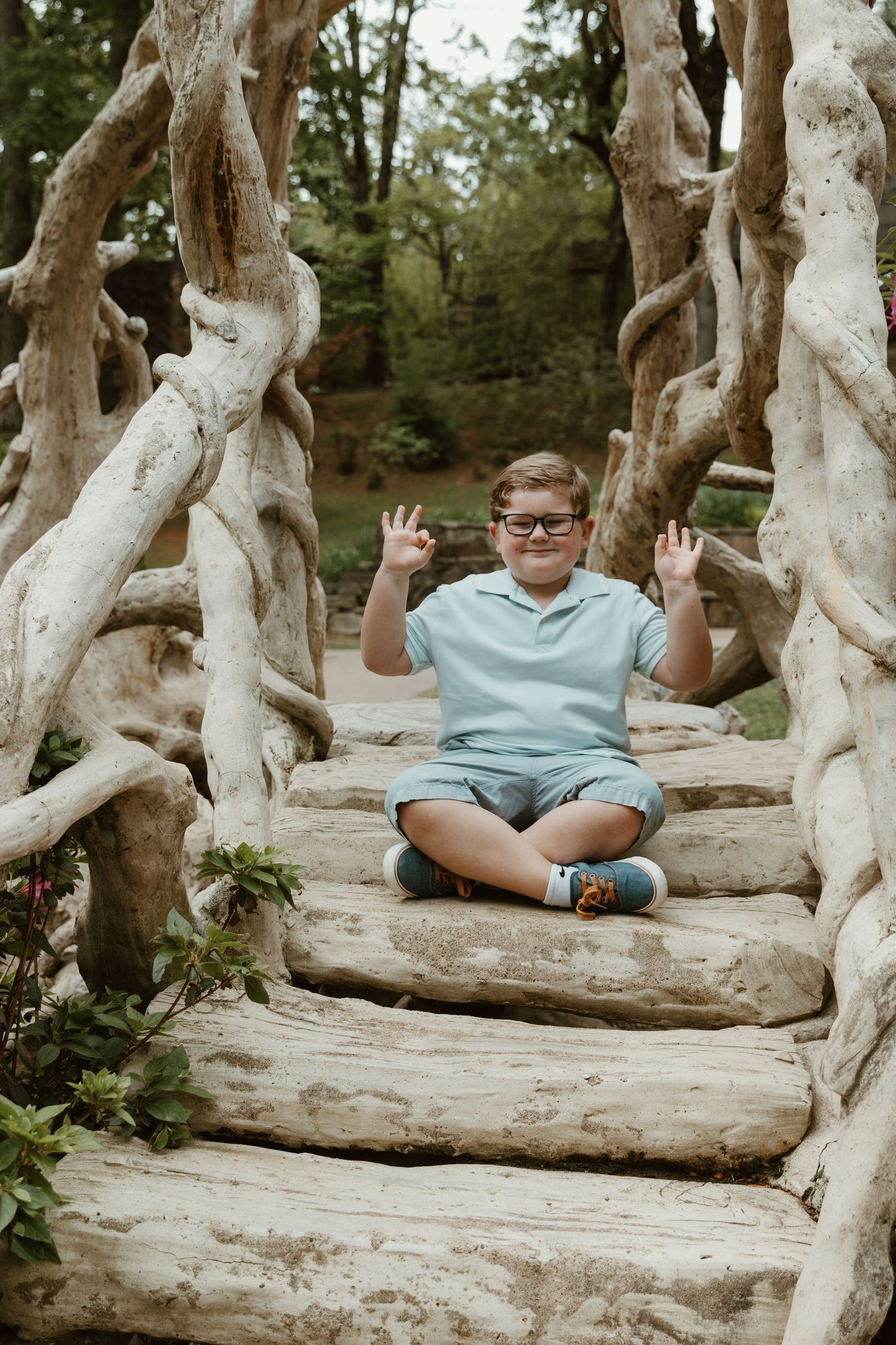 A child in glasses sits playfully on rustic wooden steps surrounded by twisted tree branches.