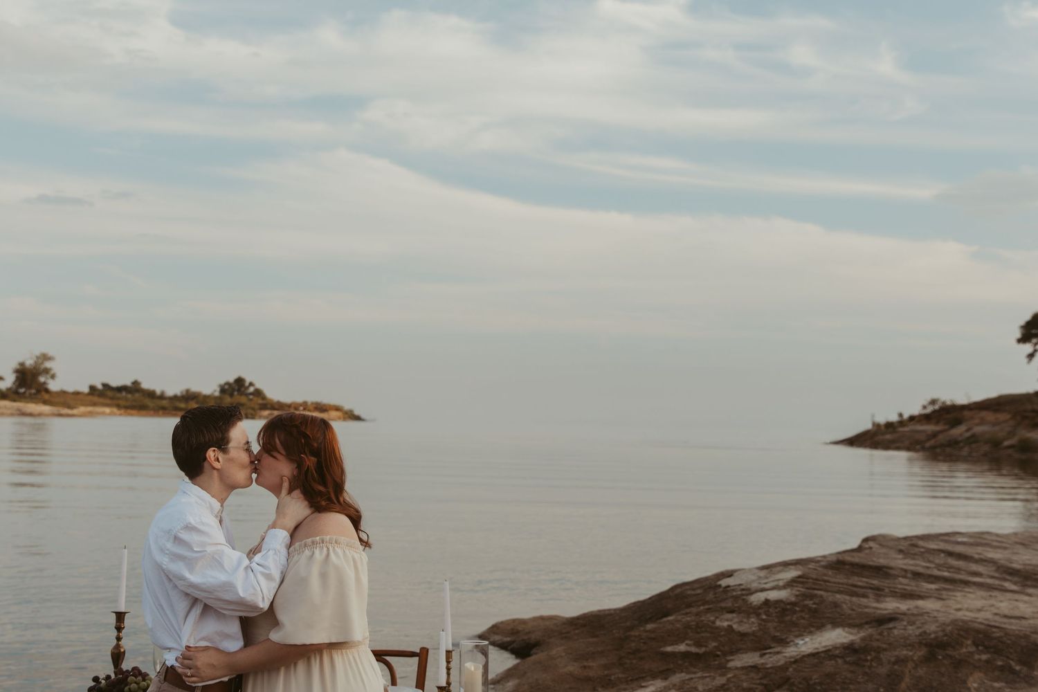 A couple in white and brown attire shares a romantic moment by the water's edge.