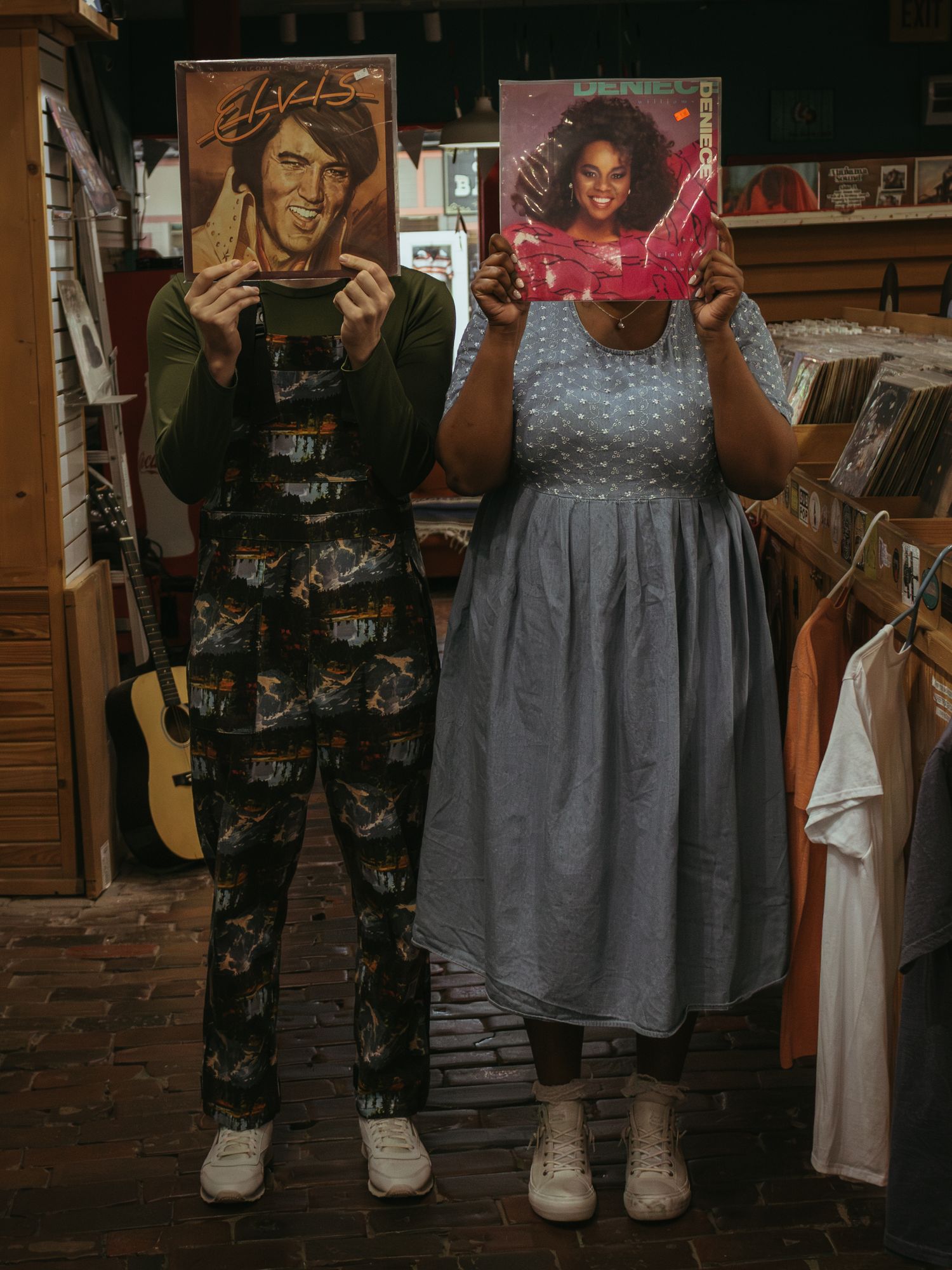 Two people holding vintage vinyl record albums in a cozy shop with wood paneling and clothing racks visible.