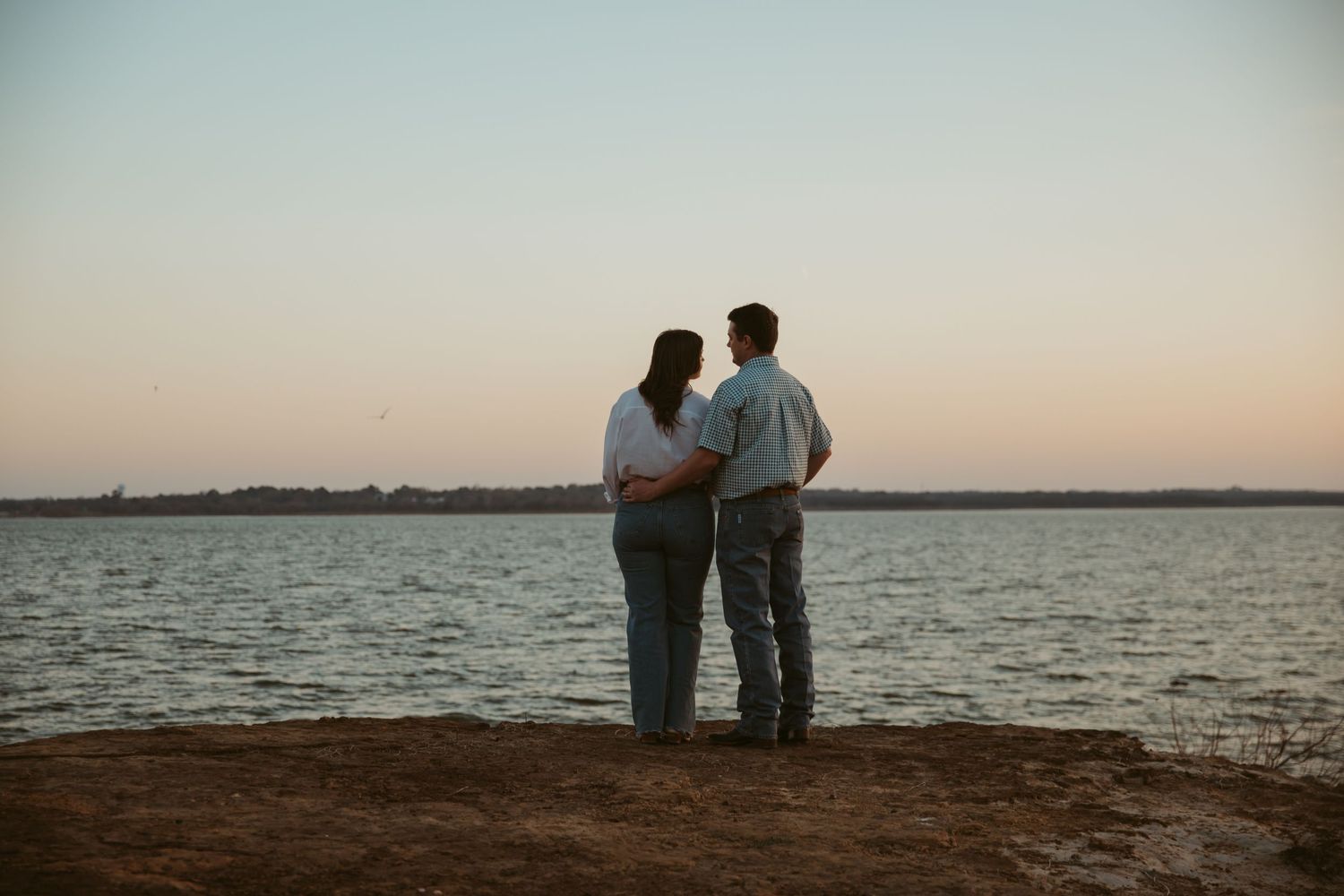Couple standing together at sunset by a calm lake shore, viewed from behind in a romantic outdoor setting.