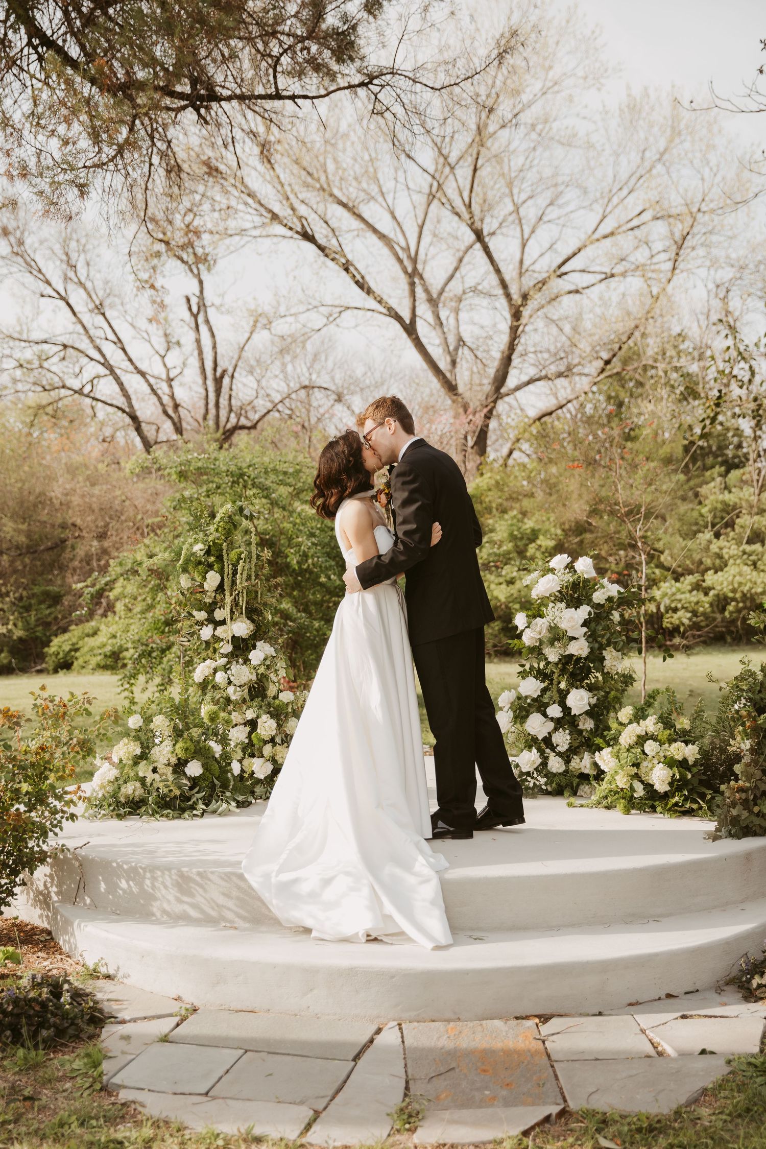 Bride and groom share romantic kiss on white circular altar surrounded by lush white floral arrangements.