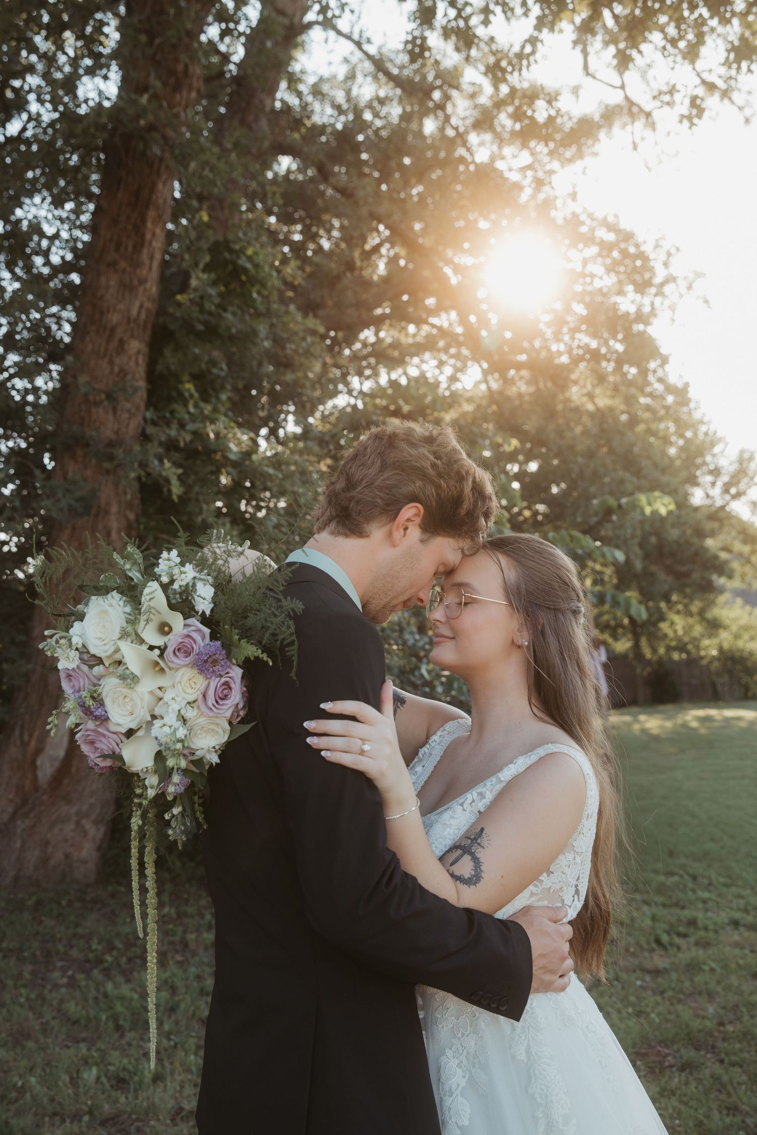 Bride and groom share romantic forehead touch outdoors at golden hour, bride holding purple and white bouquet.