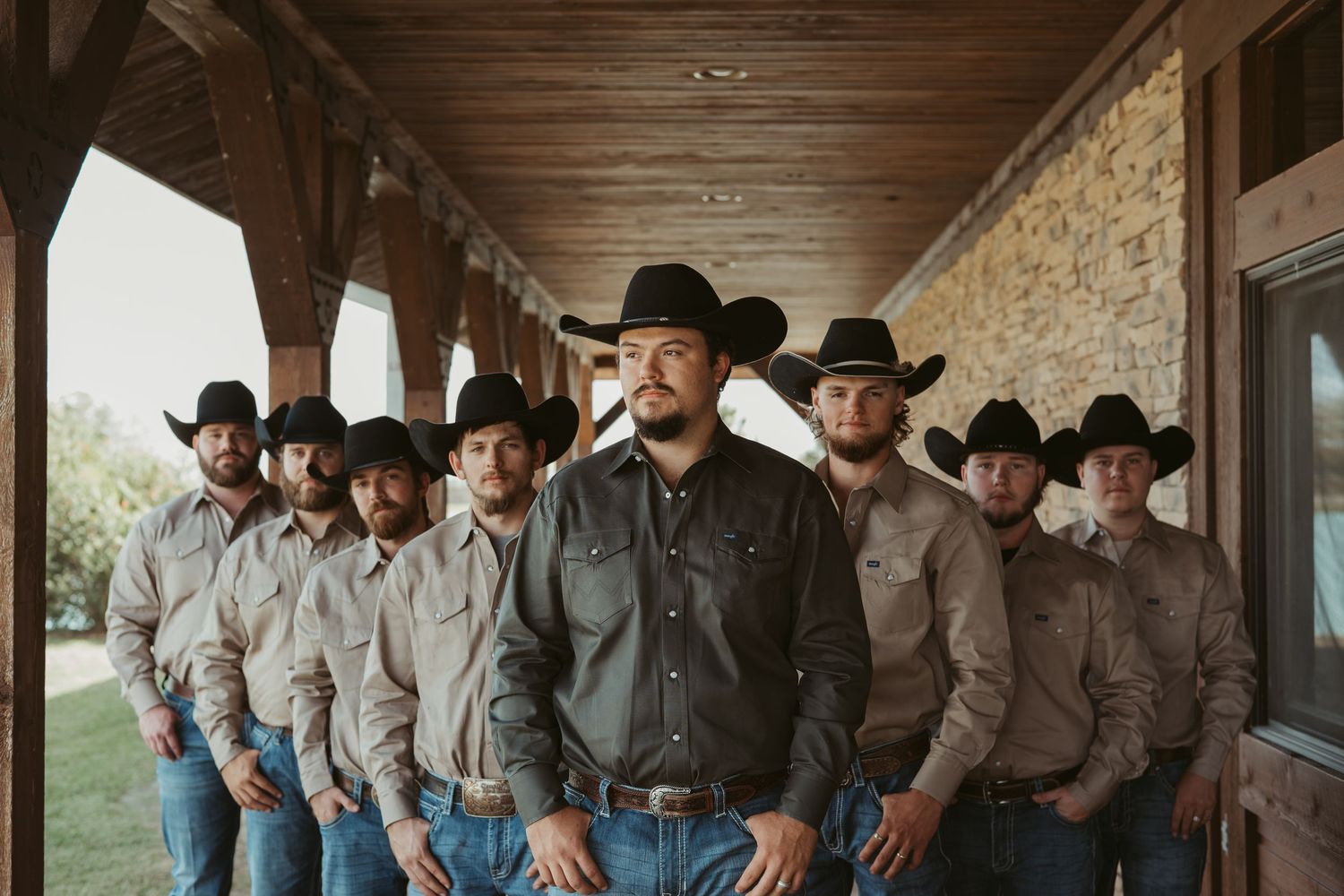 A line of cowboys in matching attire and black hats standing against a rustic wooden porch. Wedding photographer DFW
