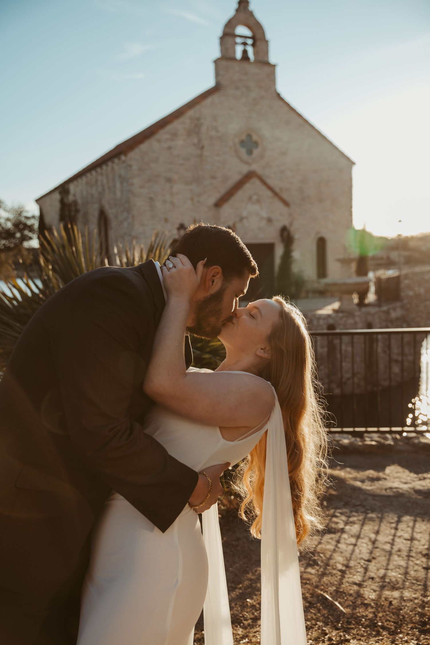 A couple shares a romantic kiss in front of a rustic stone church at sunset.