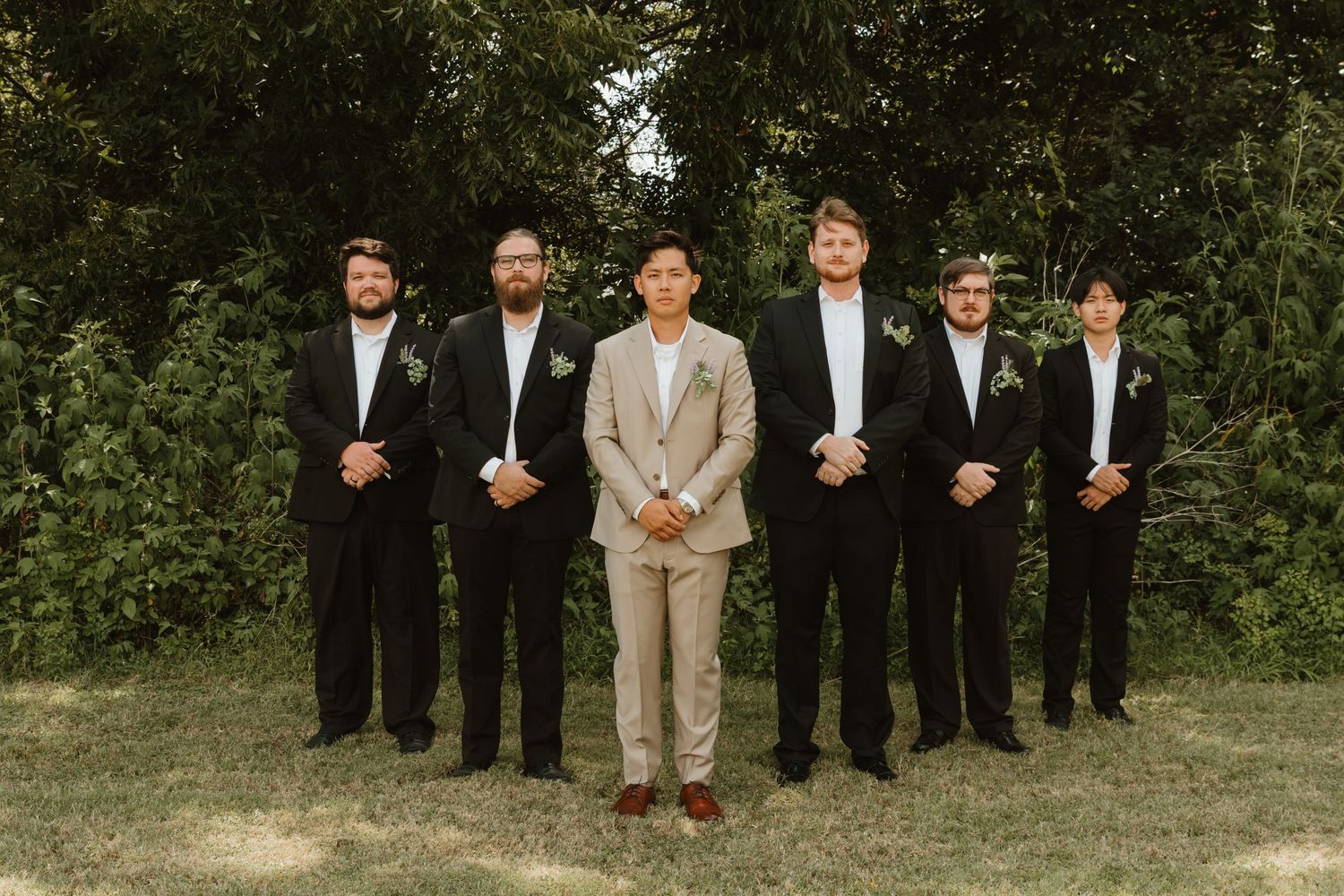Wedding party poses outdoors with groomsmen in black suits and one in light gray.