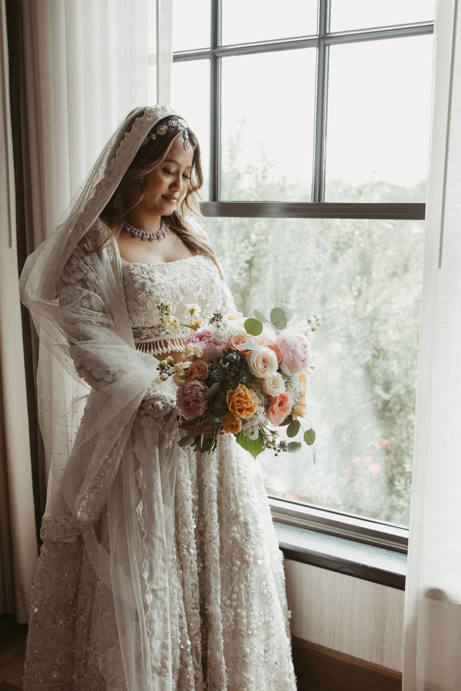 A bride in a vintage lace wedding dress holds a beautiful bouquet of pink and peach flowers by a sunlit window.