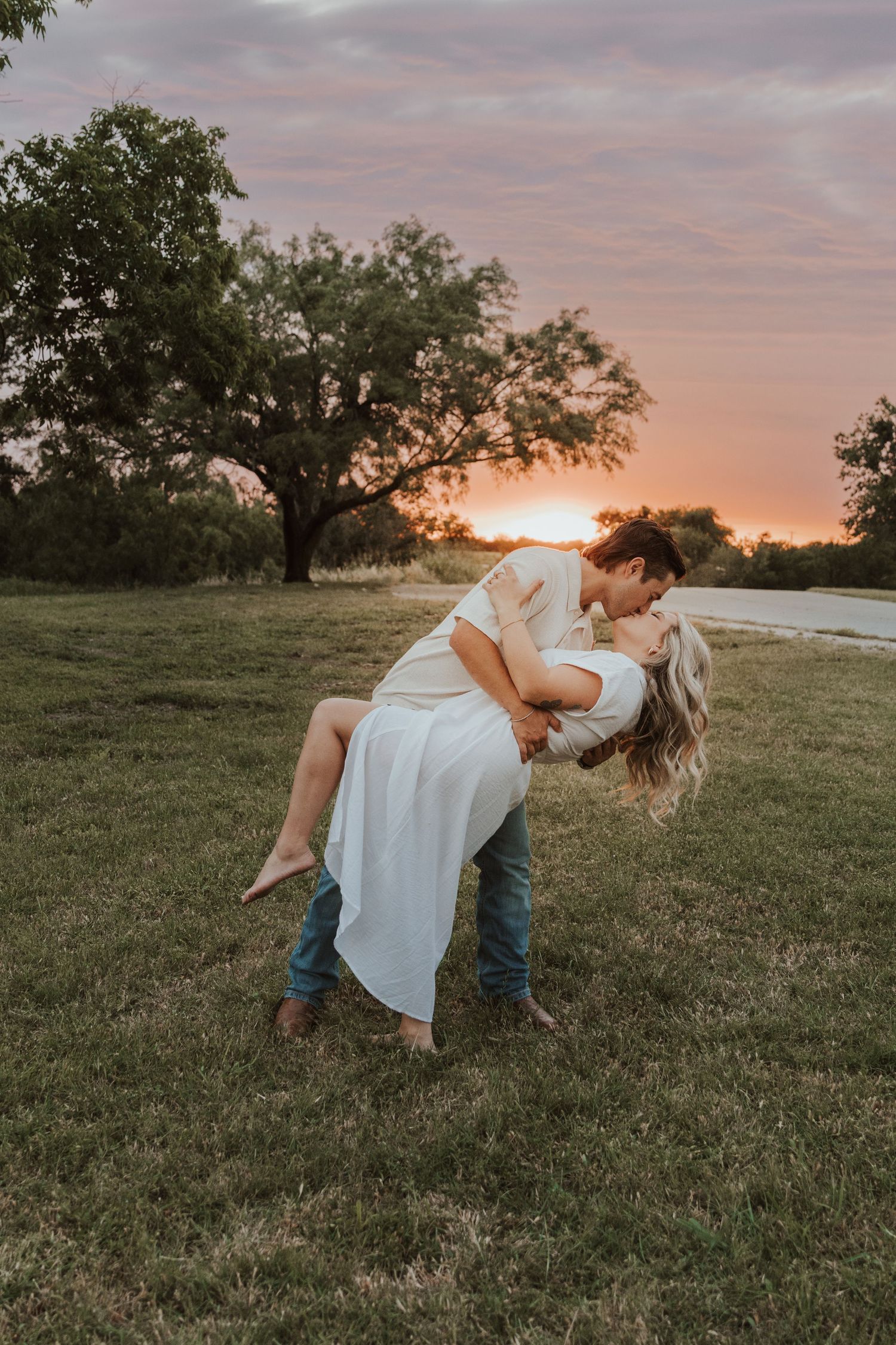 Romantic couple embraces in a dip pose against a dreamy sunset background with trees and grass.