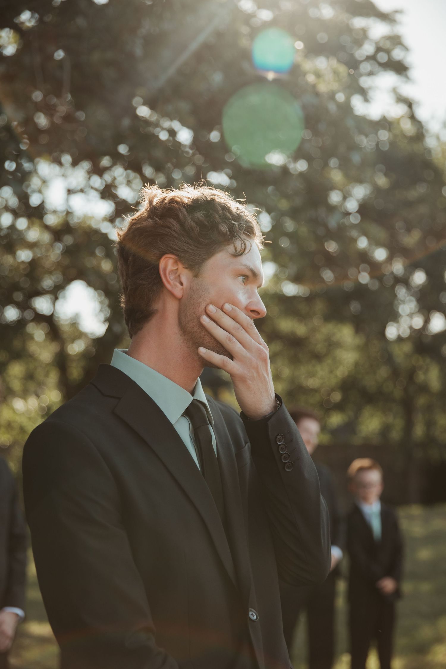 Emotional groom in dark suit covering his mouth as bride approaches during outdoor wedding ceremony.
