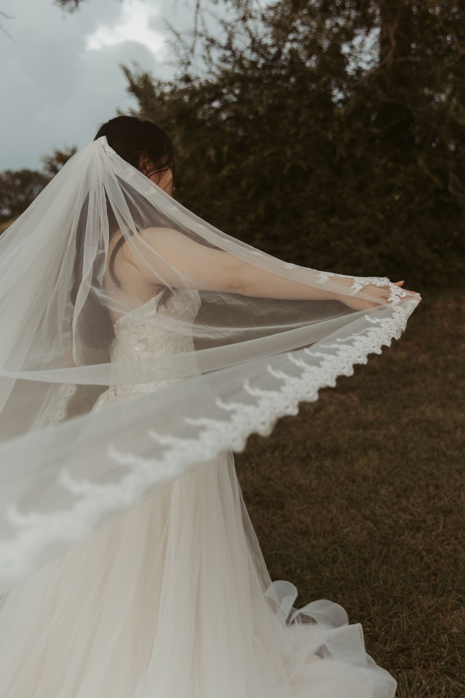 A flowing cathedral length wedding veil with lace trim billows gracefully in the wind during an outdoor bridal photoshoot.