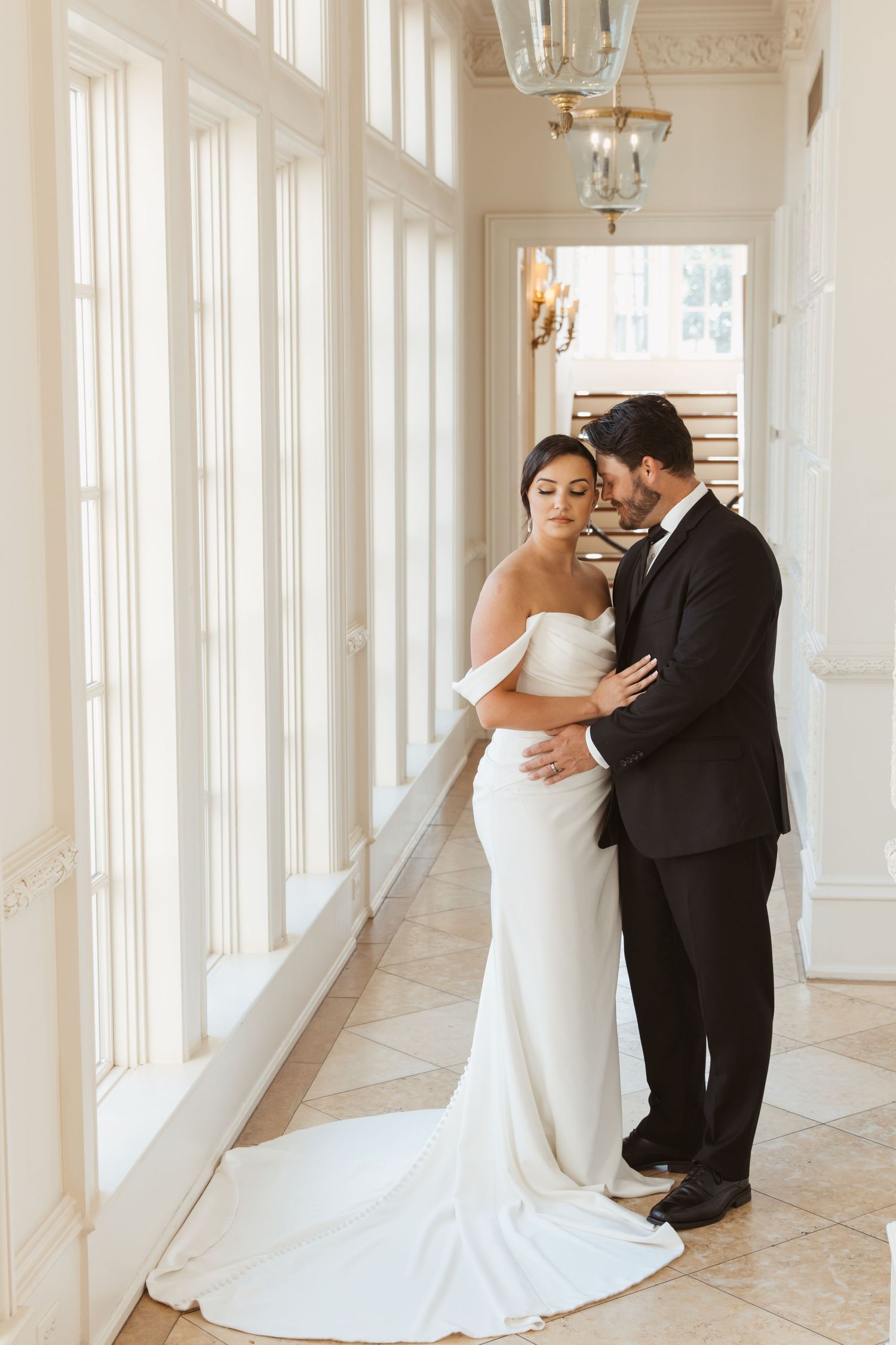 Wedding couple shares intimate moment in bright sunlit hallway with white marble floors and classical decor.