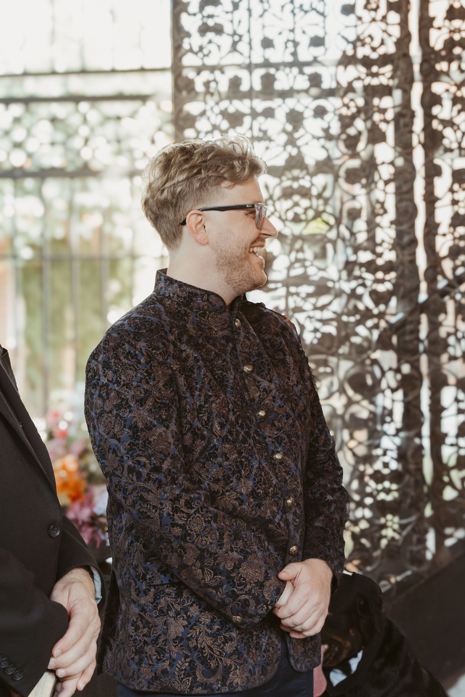 A person wearing glasses and a dark patterned button-up shirt stands in front of a decorative backdrop at a wedding venue.