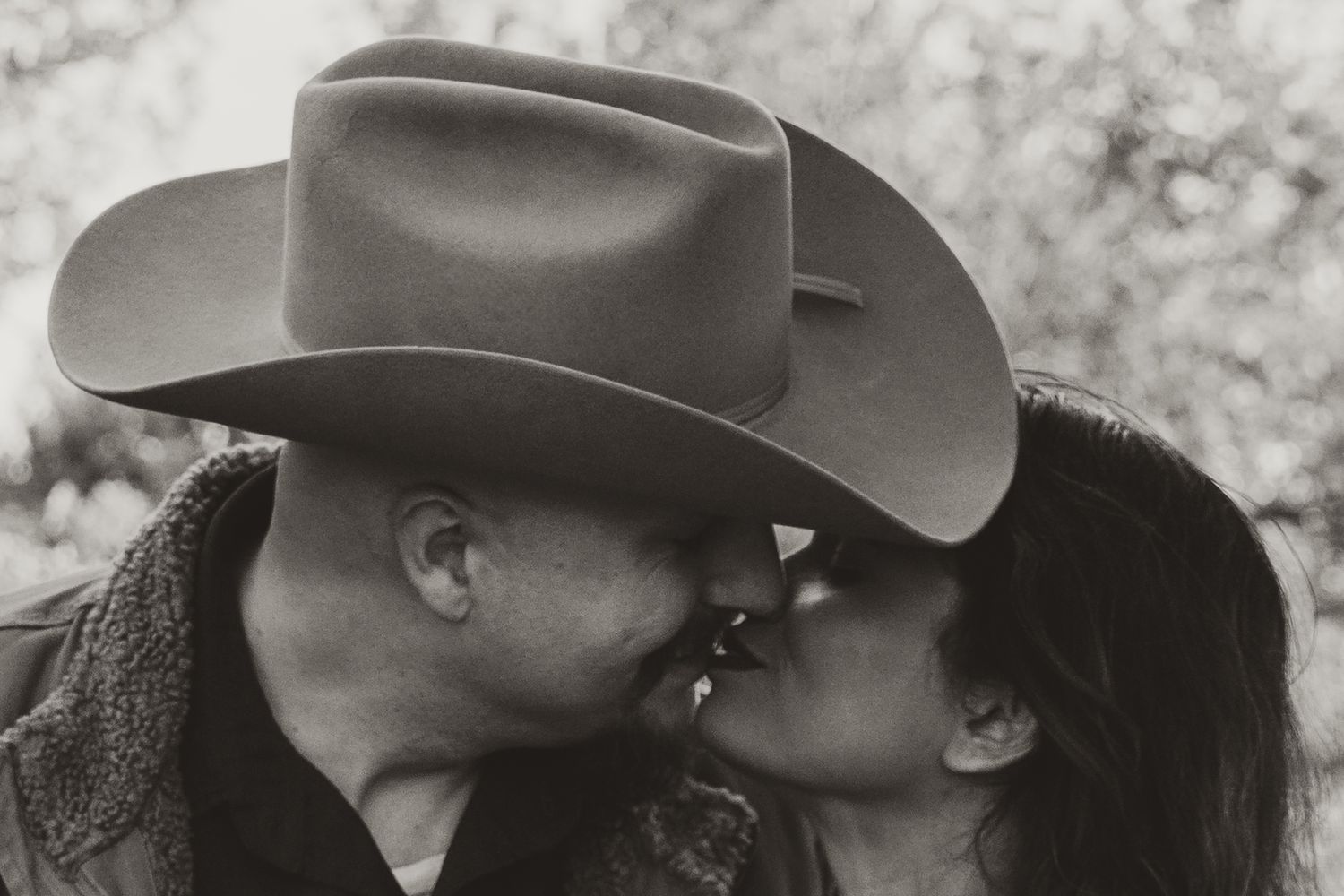 A romantic black and white photo of a cowboy hat-wearing person sharing an intimate kiss with their partner.