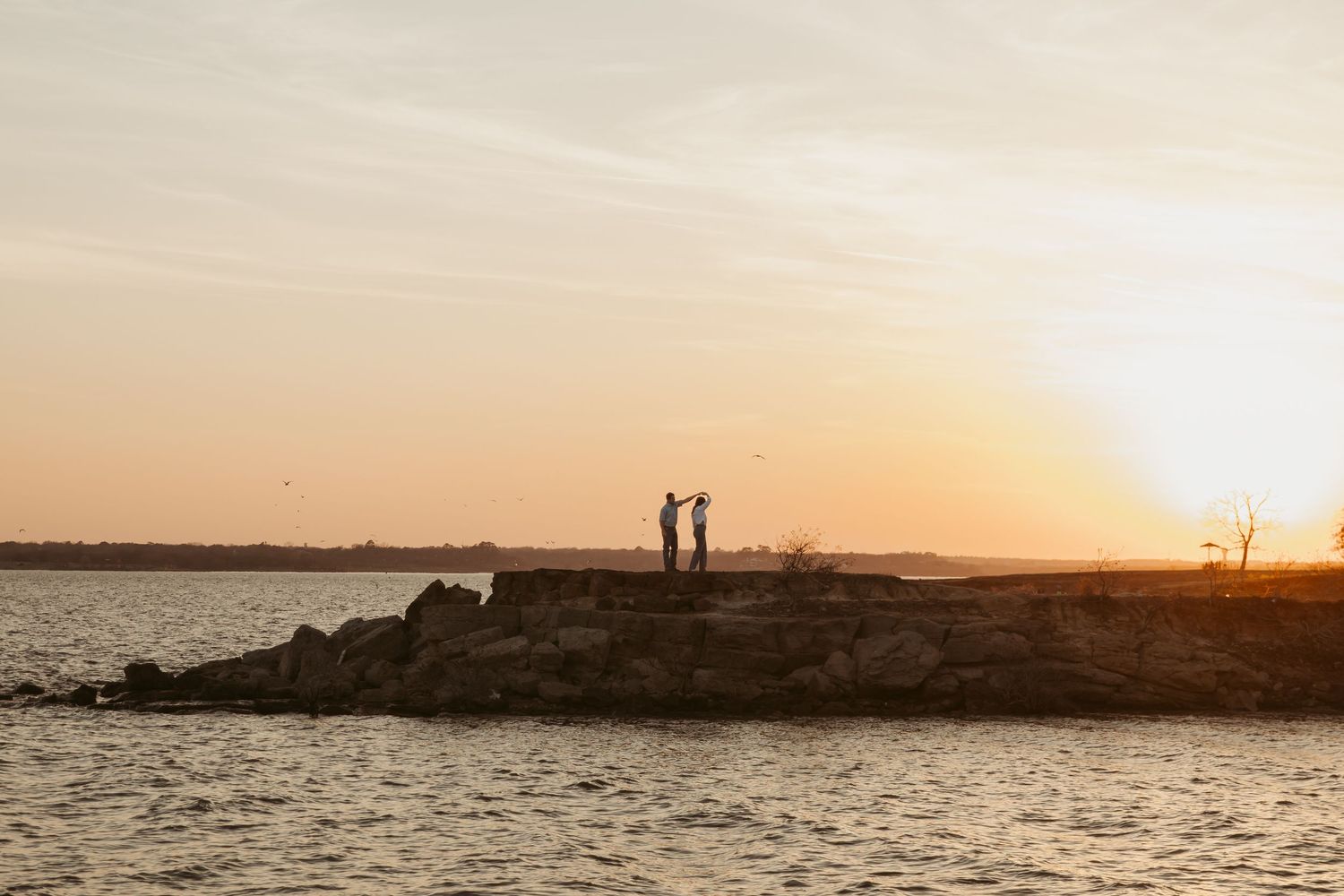 Two silhouetted figures stand on rocky shoreline at golden sunset over calm water.