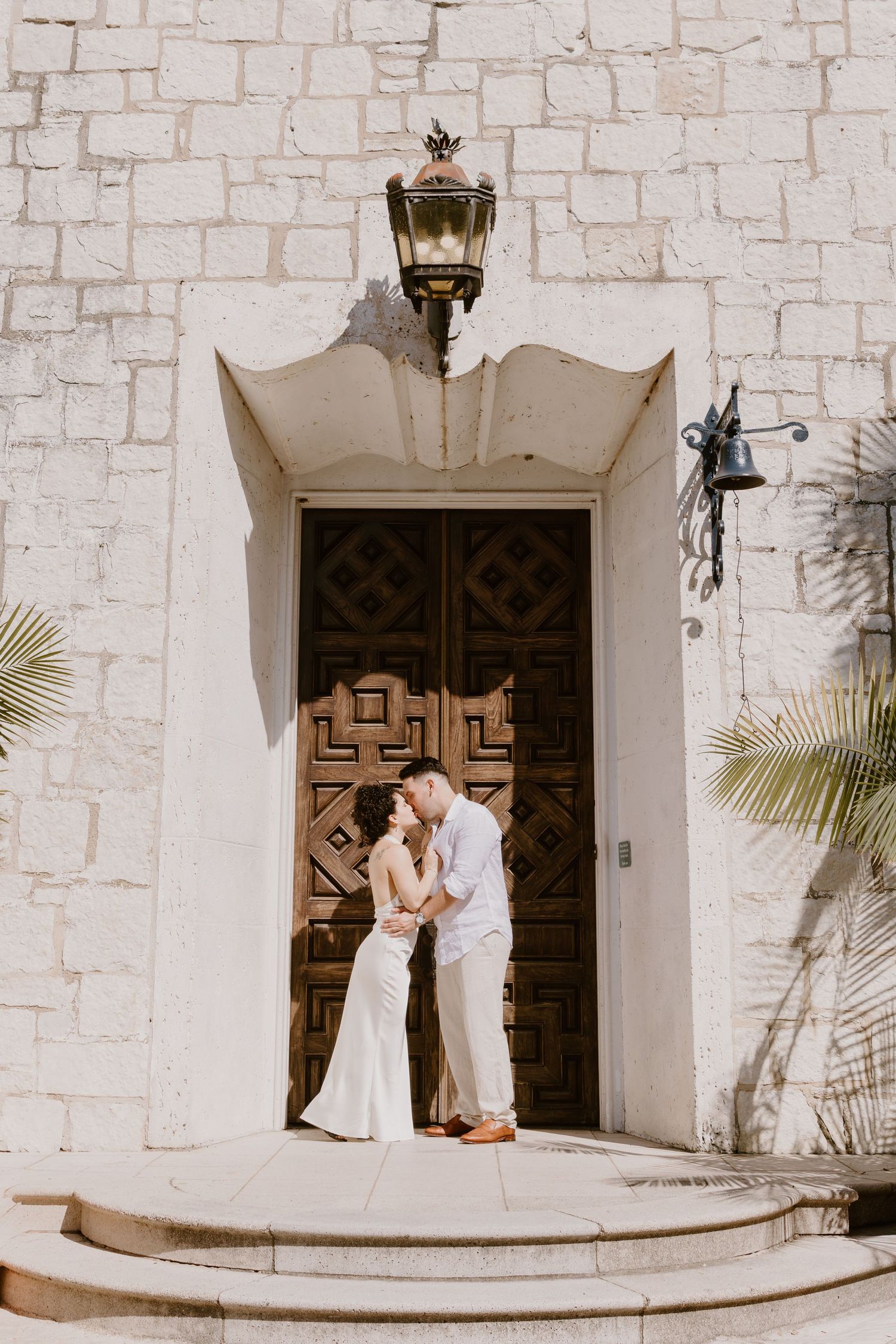 Elegant couple in white poses in front of a Spanish colonial church with ornate wooden doors.