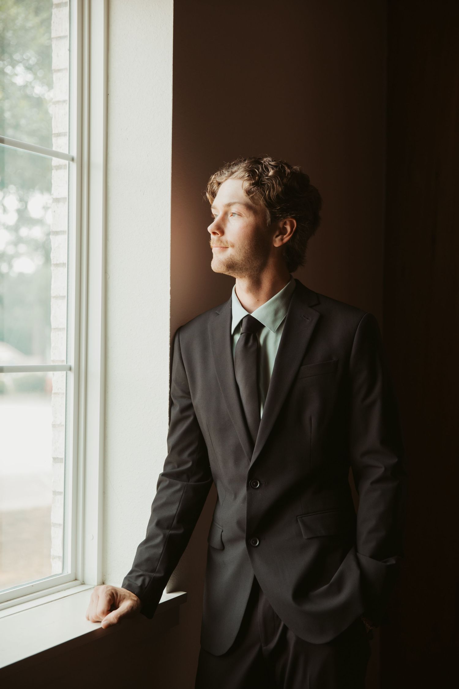 Young man in dark suit and tie gazing out a window with natural light, leaning against a dark wall.