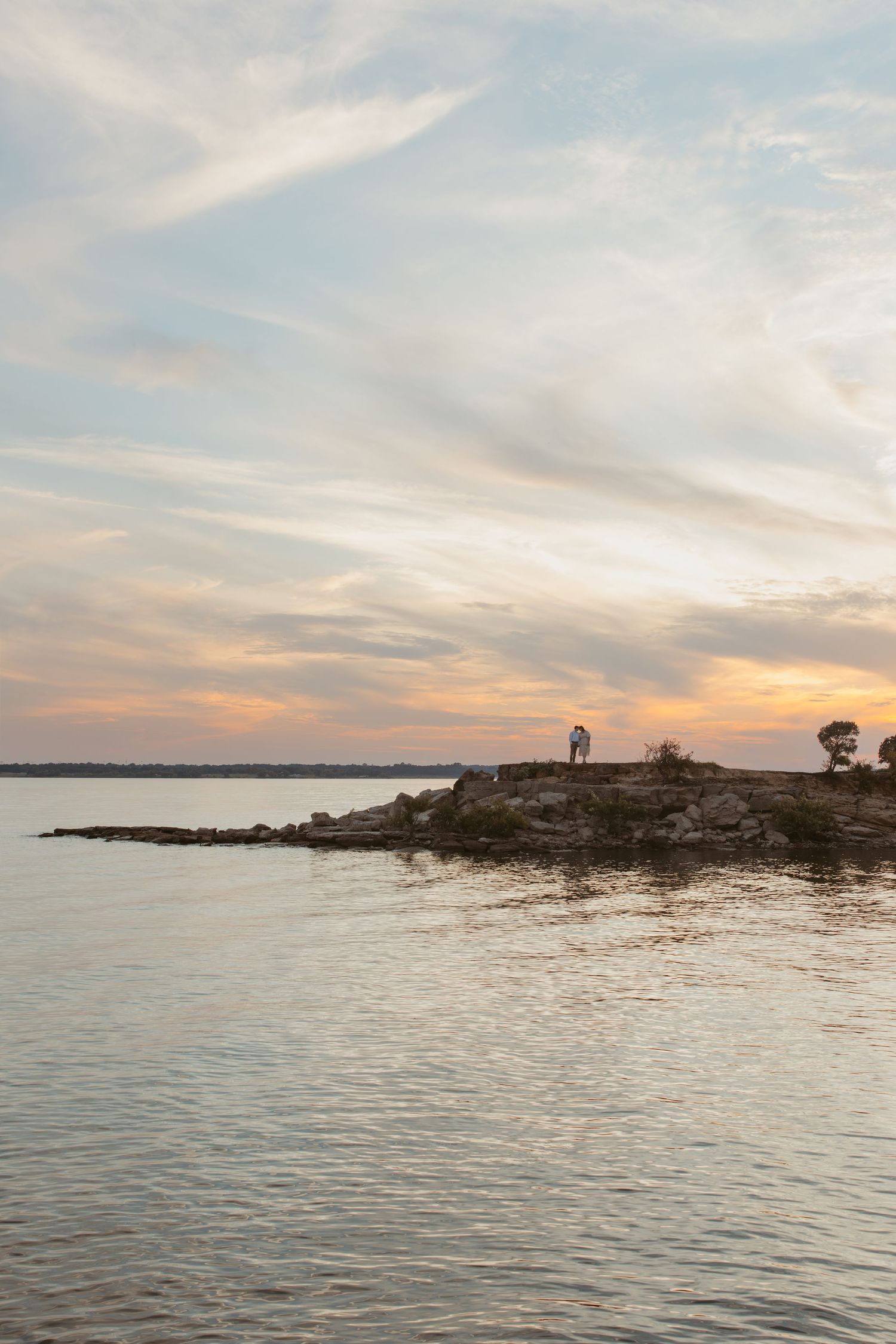 Scenic panoramic view of a tranquil lake at sunset with silhouetted rocky shoreline and warm orange sky.