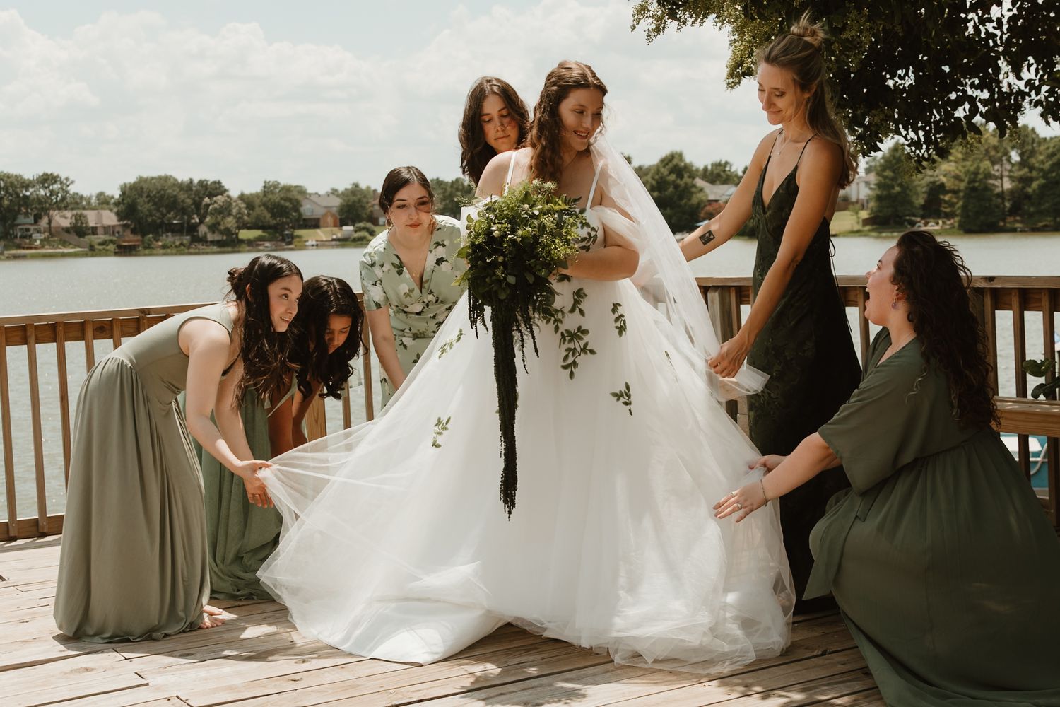 A wedding party in sage green dresses helps adjust a flowing white gown on an outdoor deck overlooking water.