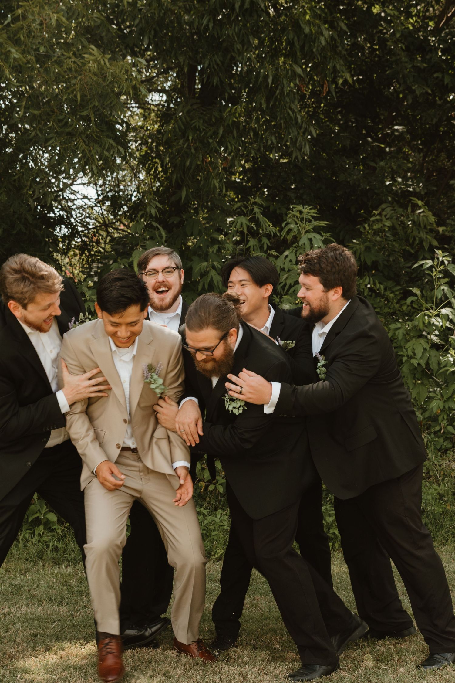 Groomsmen in black and beige suits laughing and posing playfully outdoors during wedding photo session.
