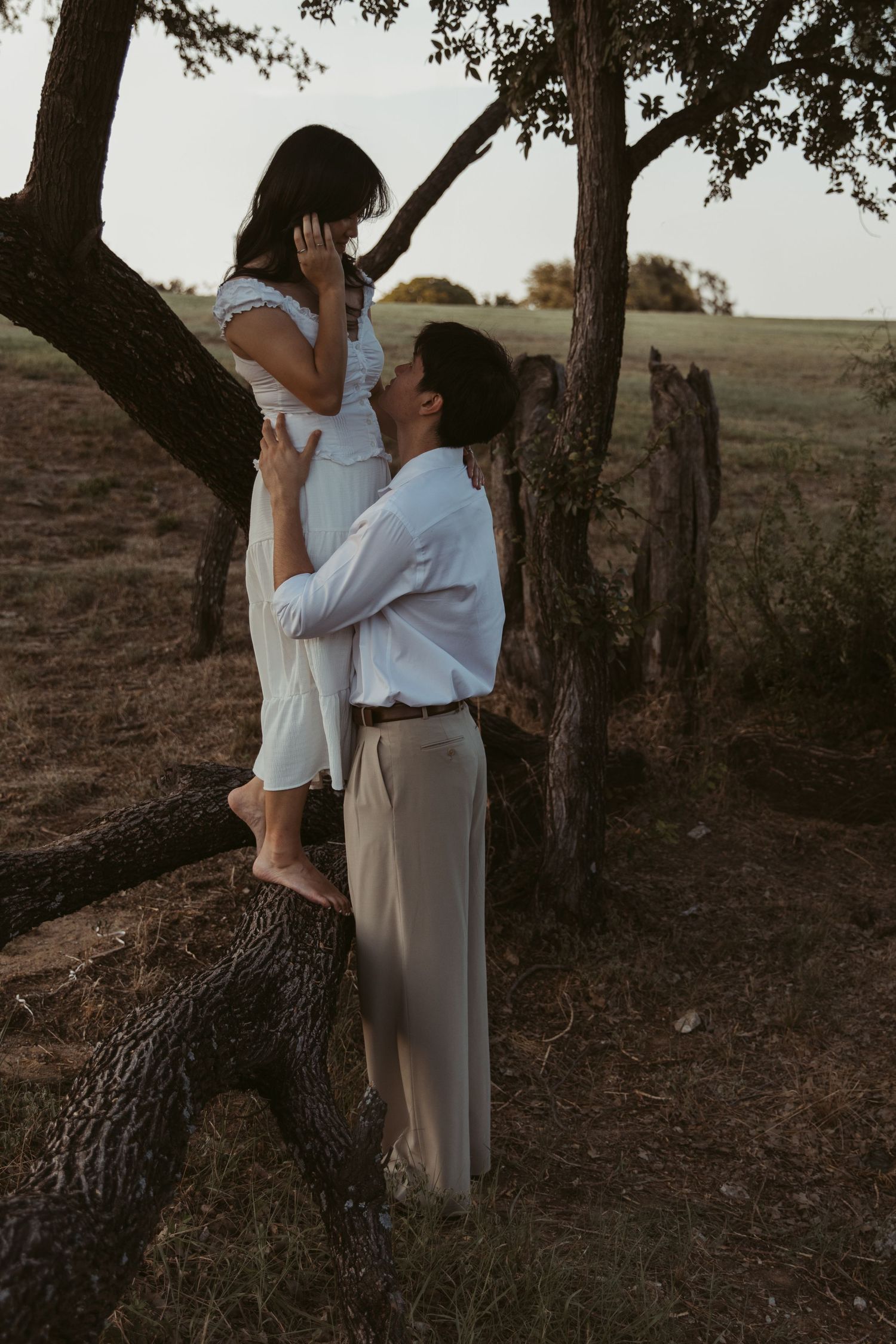Couple shares romantic moment under tree branches at sunset in rural setting.