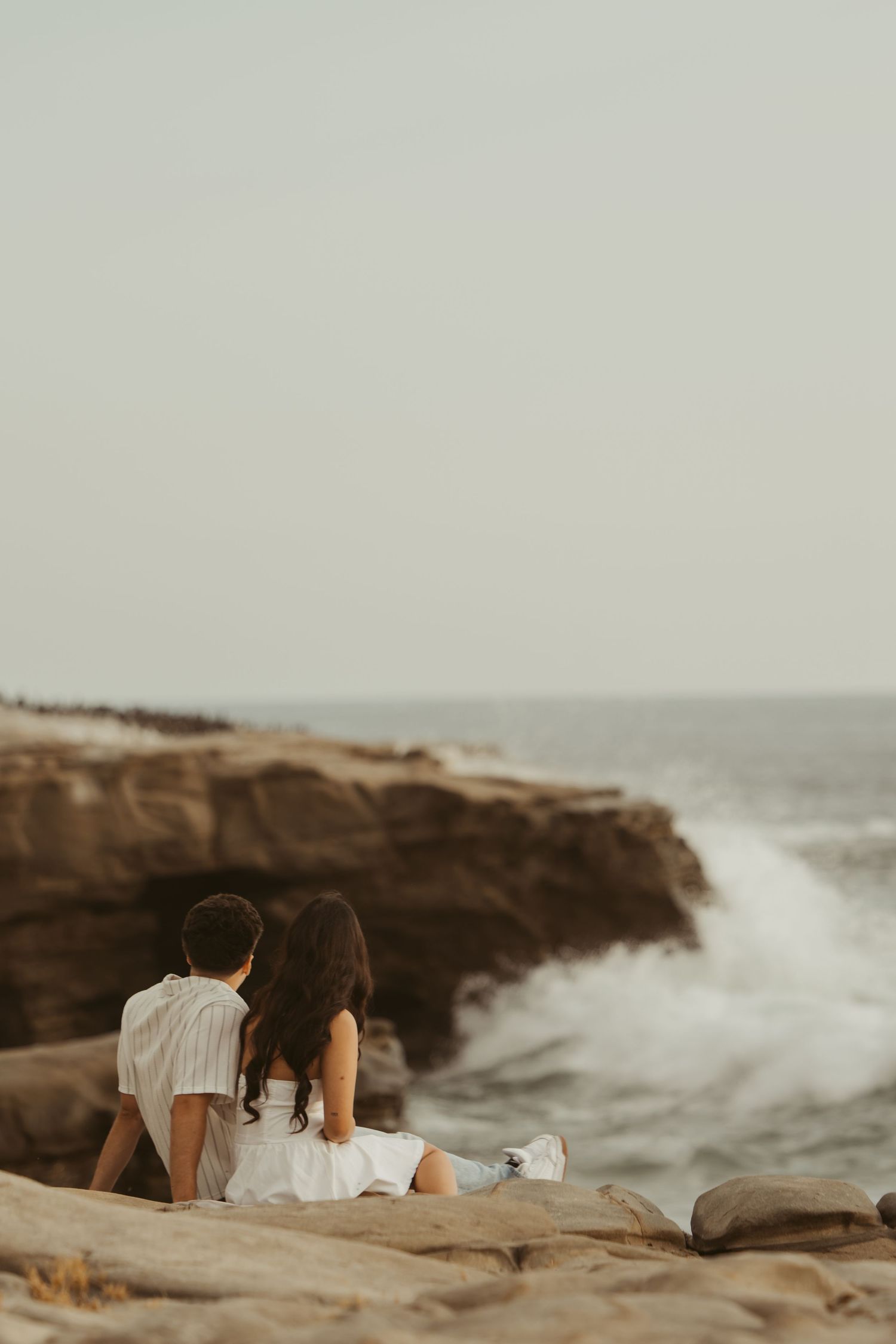 Couple sitting on rocky coastal cliffs watching dramatic ocean waves crash, both dressed in white, viewed from behind.