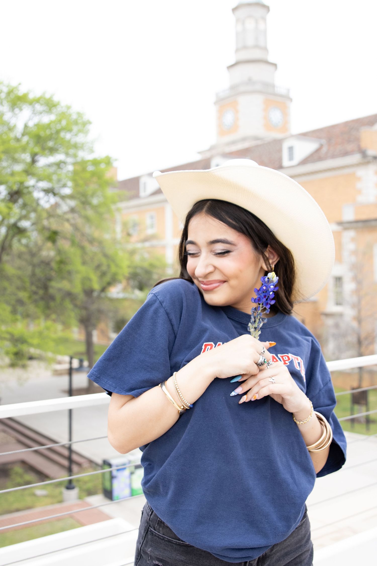 Someone in a navy blue t-shirt, white cowboy hat and statement blue earrings poses with closed eyes against an institutional building.