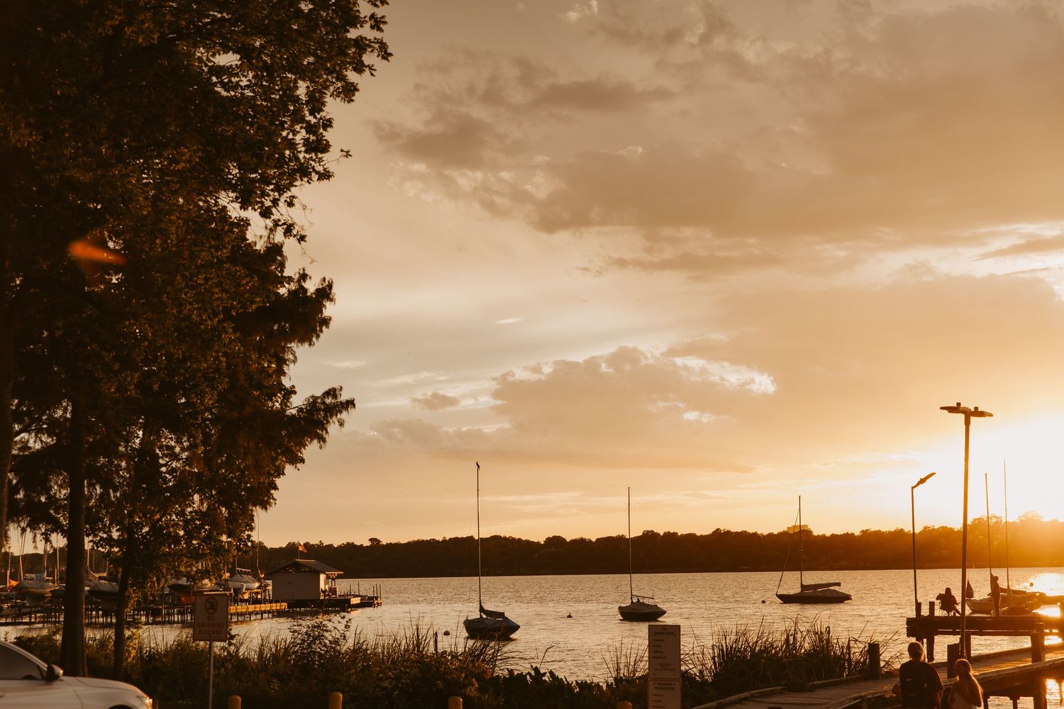 Golden sunset over a calm harbor with sailboats on the water, framed by silhouetted trees and a glowing sky.