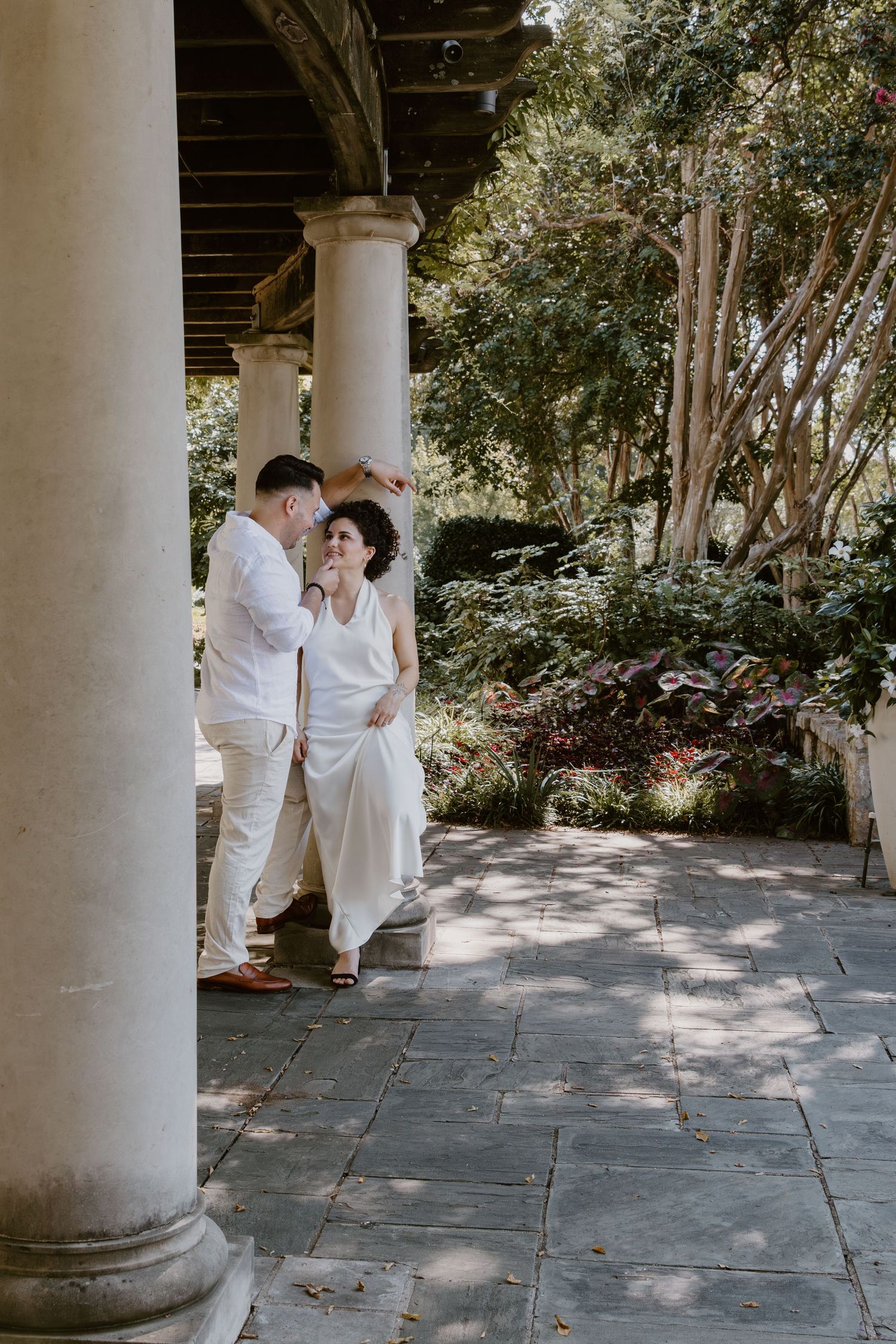 A couple in matching white outfits shares an intimate moment under a columned portico.