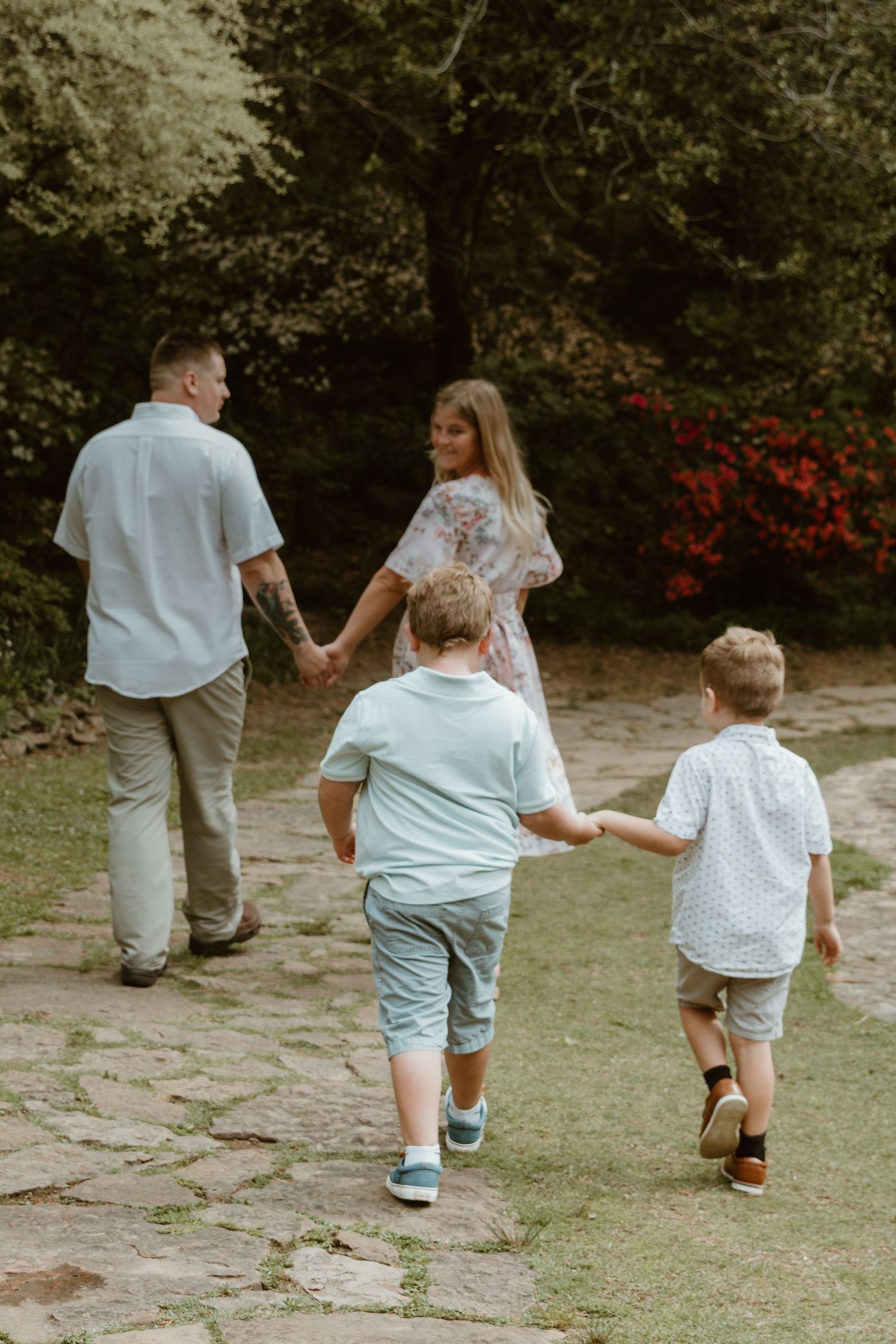 A family walks together holding hands on a stone path in a garden with blooming red flowers in the background.