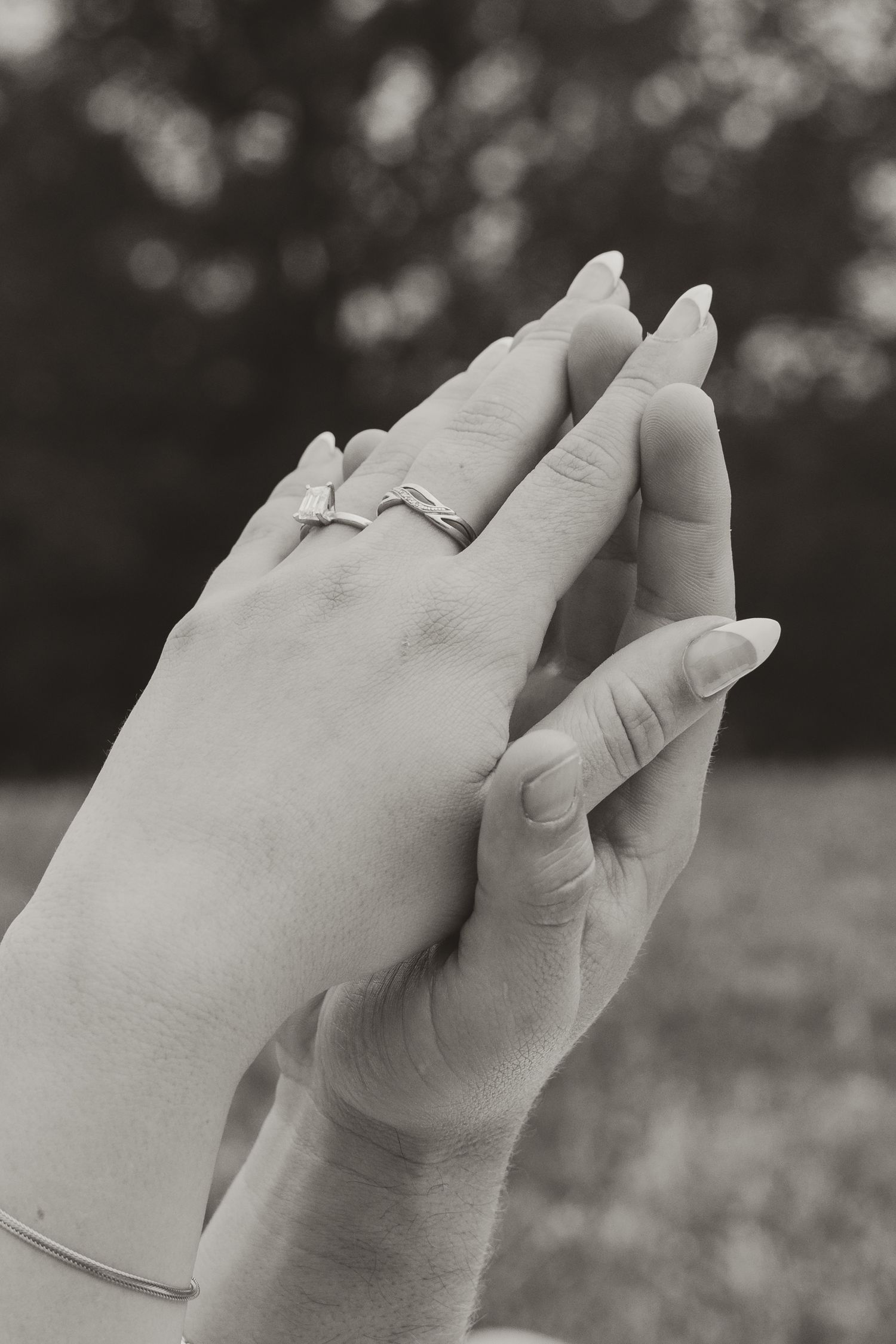Black and white photograph of hands clasped together showing an engagement ring against a blurred outdoor background.