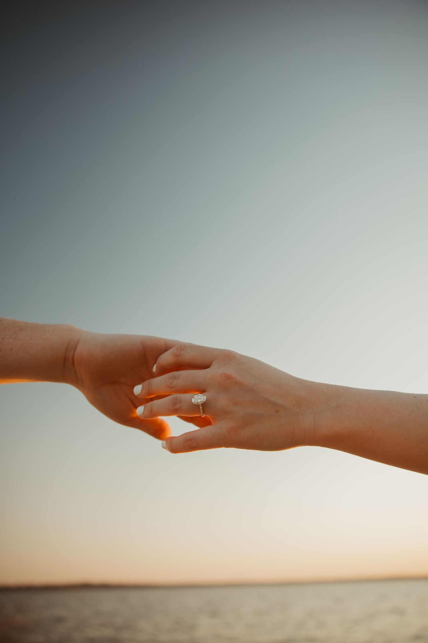 Two hands gently touching at sunset by the ocean, with an engagement ring visible on the womans finger.