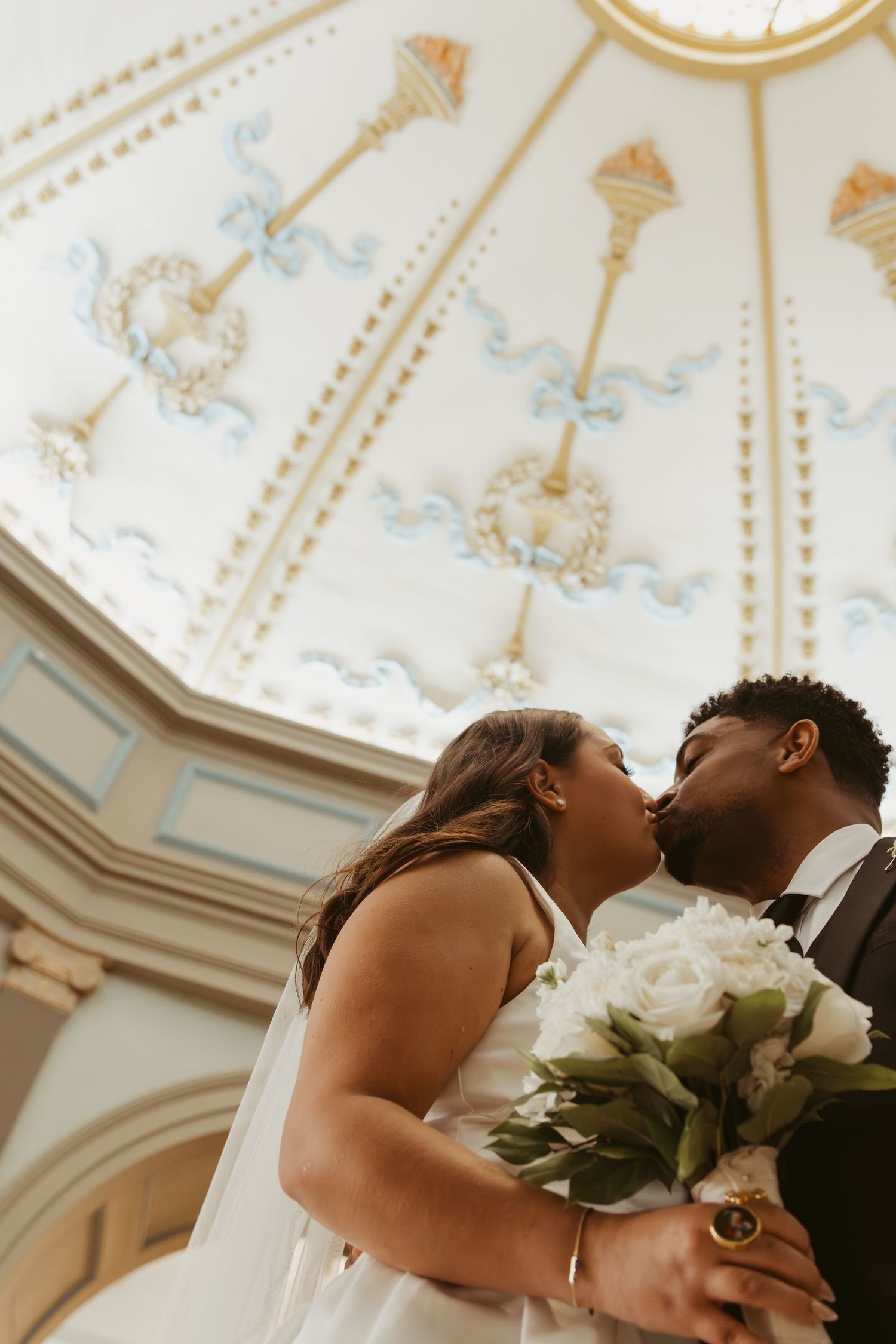 A couple shares a romantic kiss during their wedding ceremony under an ornate white ceiling with decorative molding. Wedding Photographer DF