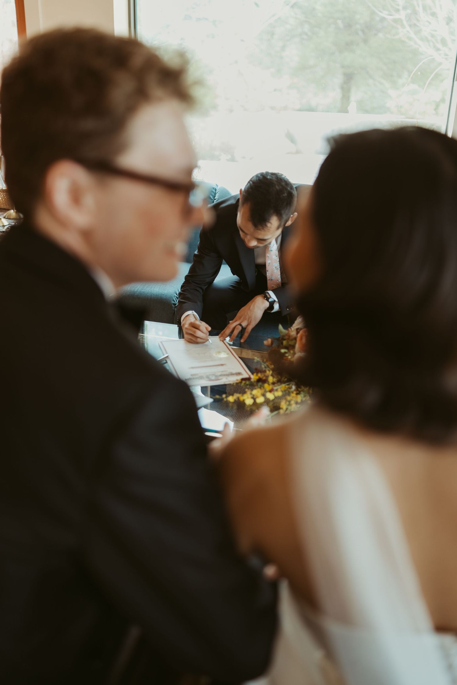 Wedding officiant signing marriage certificate while bride and groom watch during intimate ceremony signing.