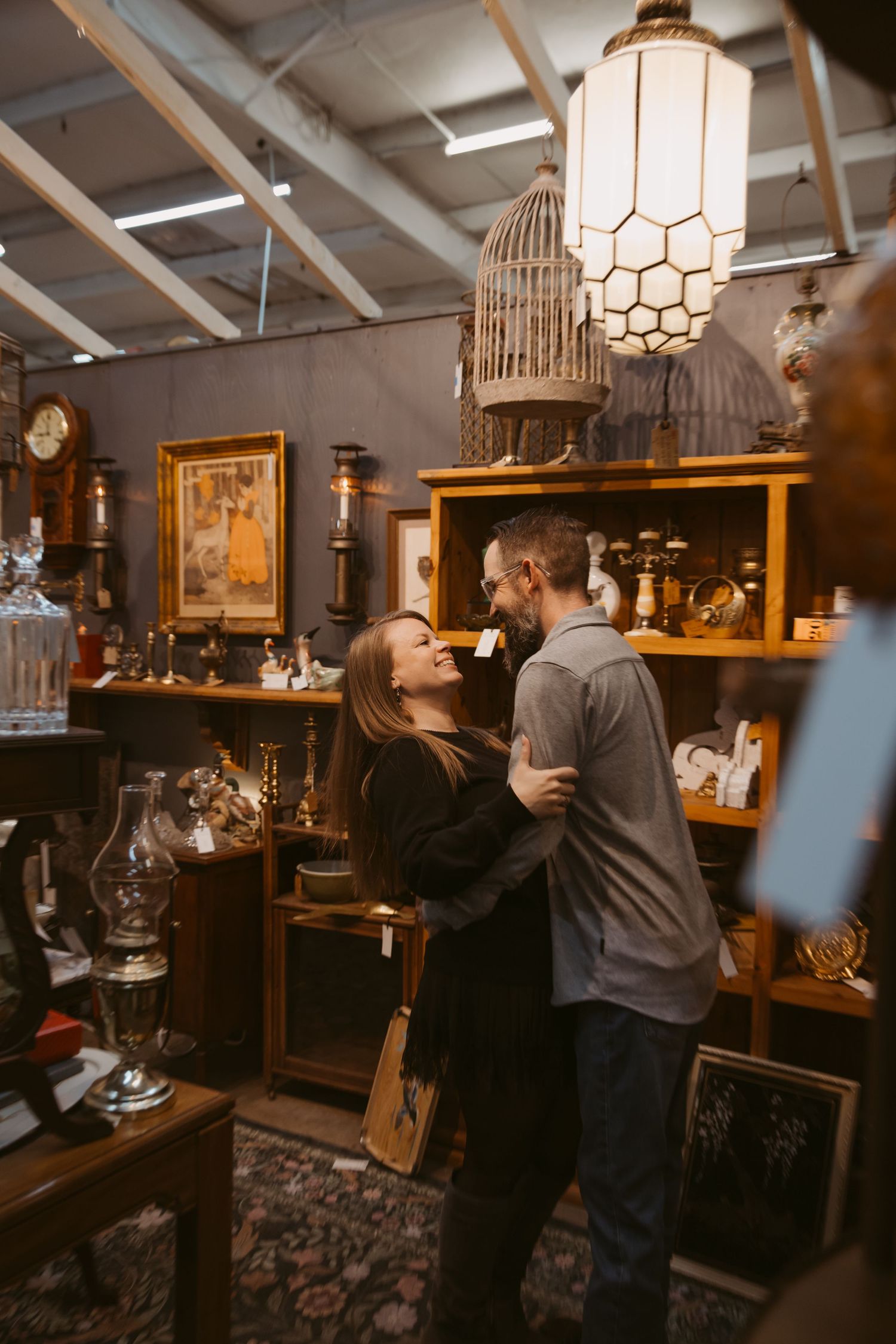 A couple embraces in a vintage store with decorative birdcages and antique furniture in warm lighting.