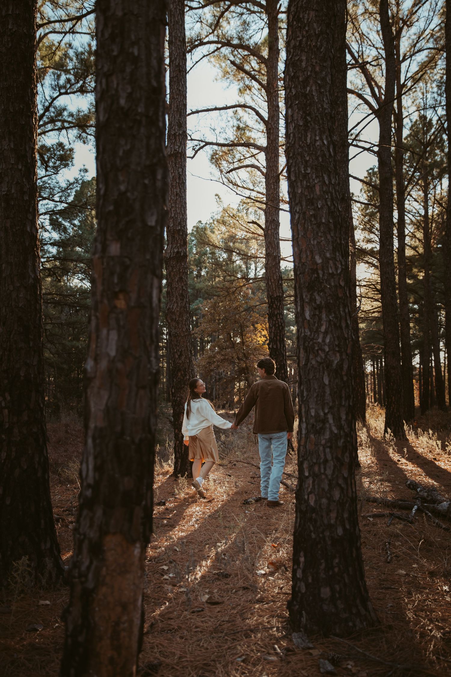 Couple walking through a sunlit pine forest holding hands on an autumn afternoon. engagement photos Fort Worth