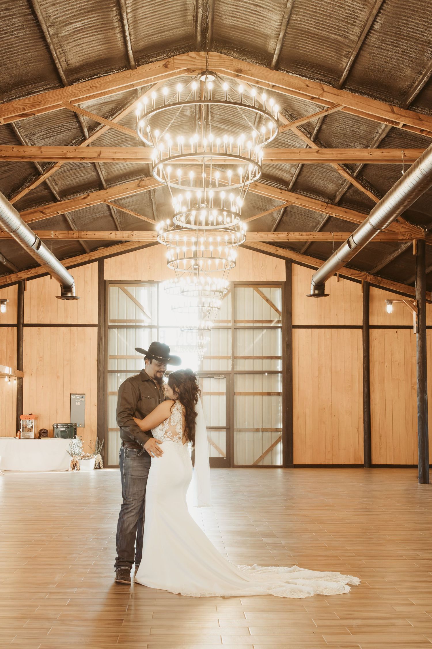 Couples dancing in a rustic barn venue with elegant circular chandeliers and wooden beams overhead. Wedding photographer DFW