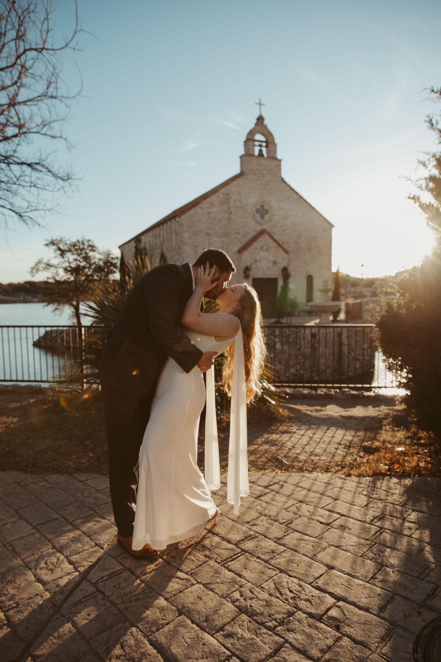 A couple in wedding attire shares an intimate moment at sunset outside a rustic stone chapel with a bell tower.