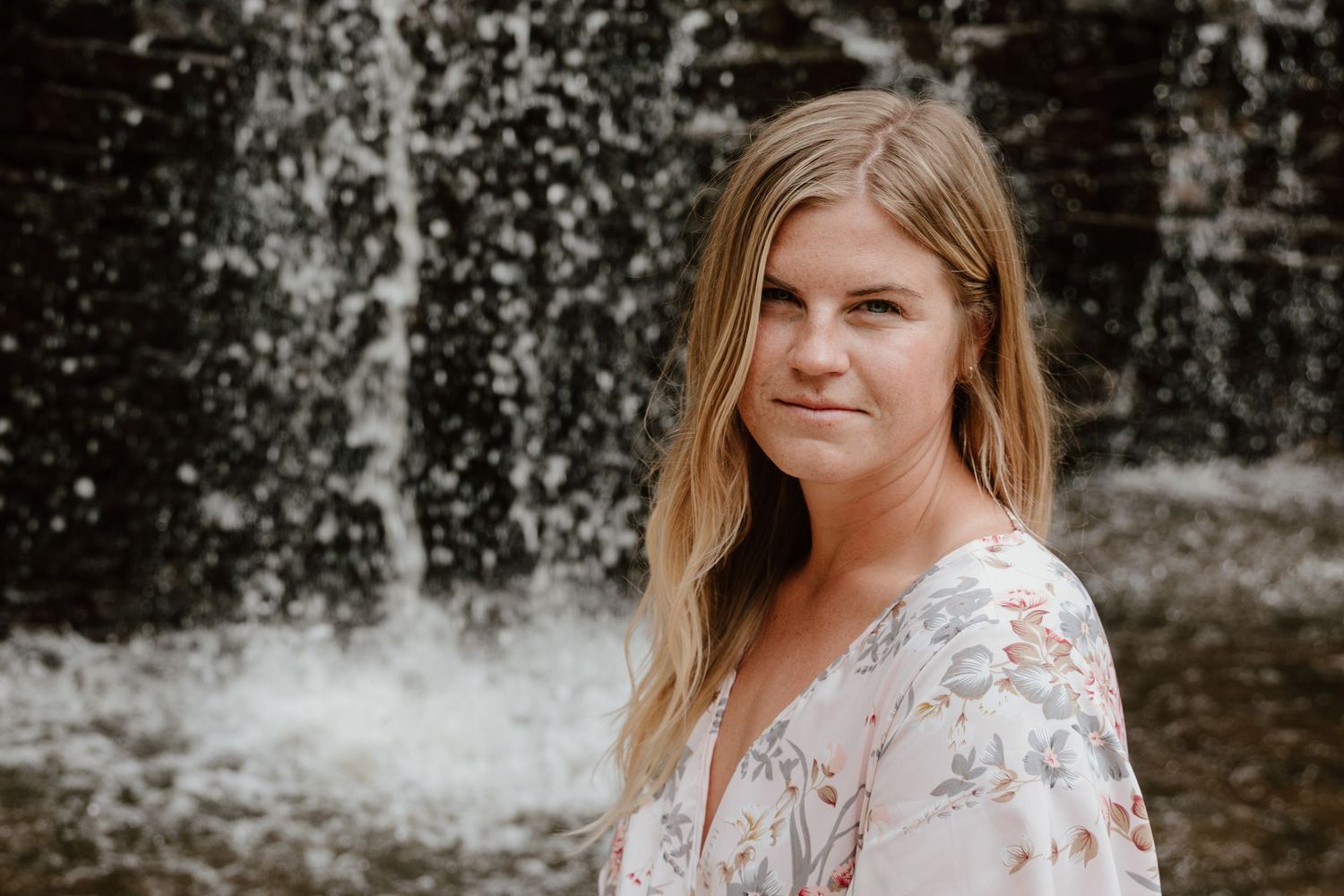 A woman in a floral dress smiles while standing in front of a cascading waterfall.