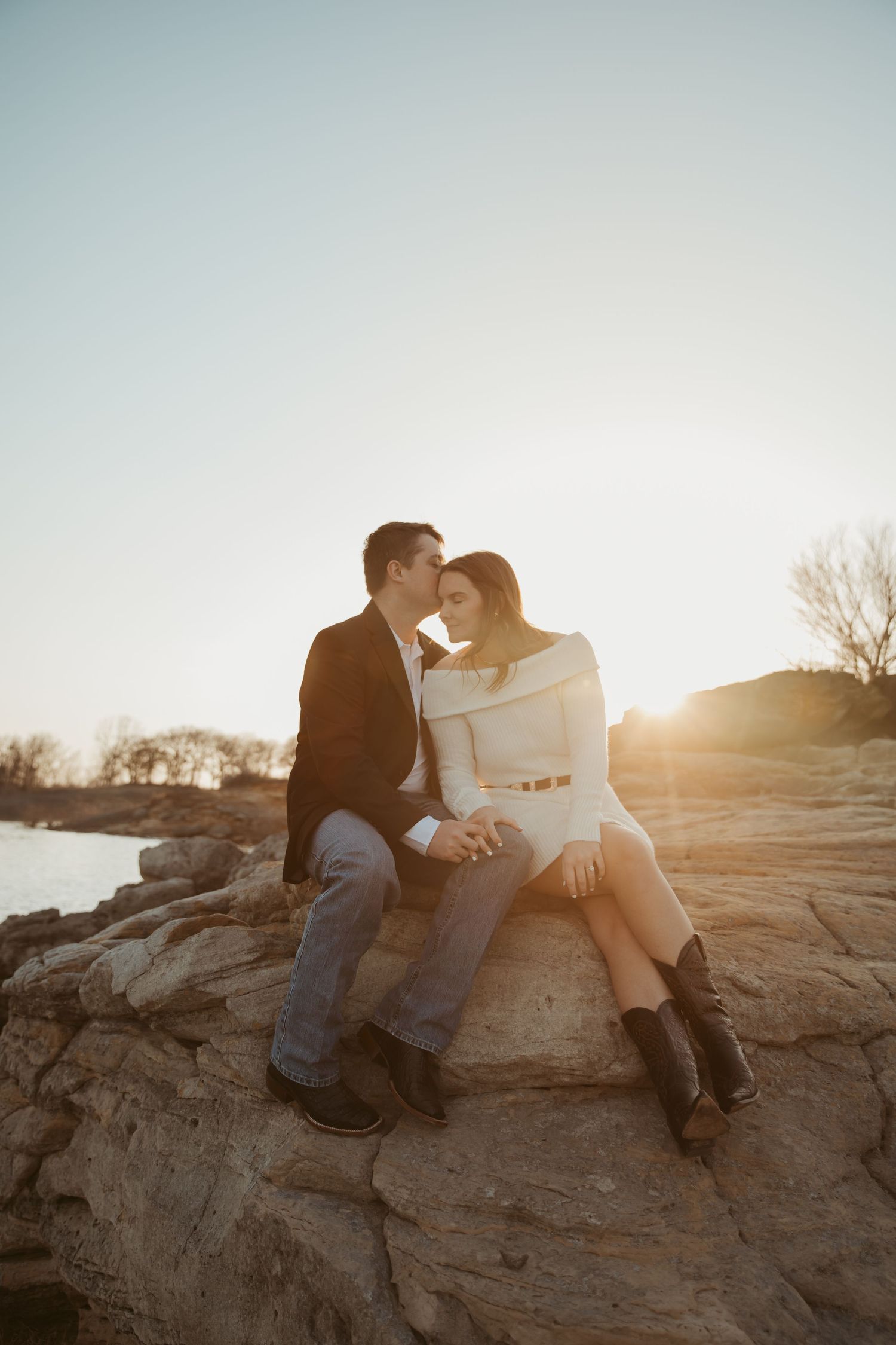 Couple sitting on rocks at sunset, man kissing woman's forehead while holding hands in romantic golden hour light.