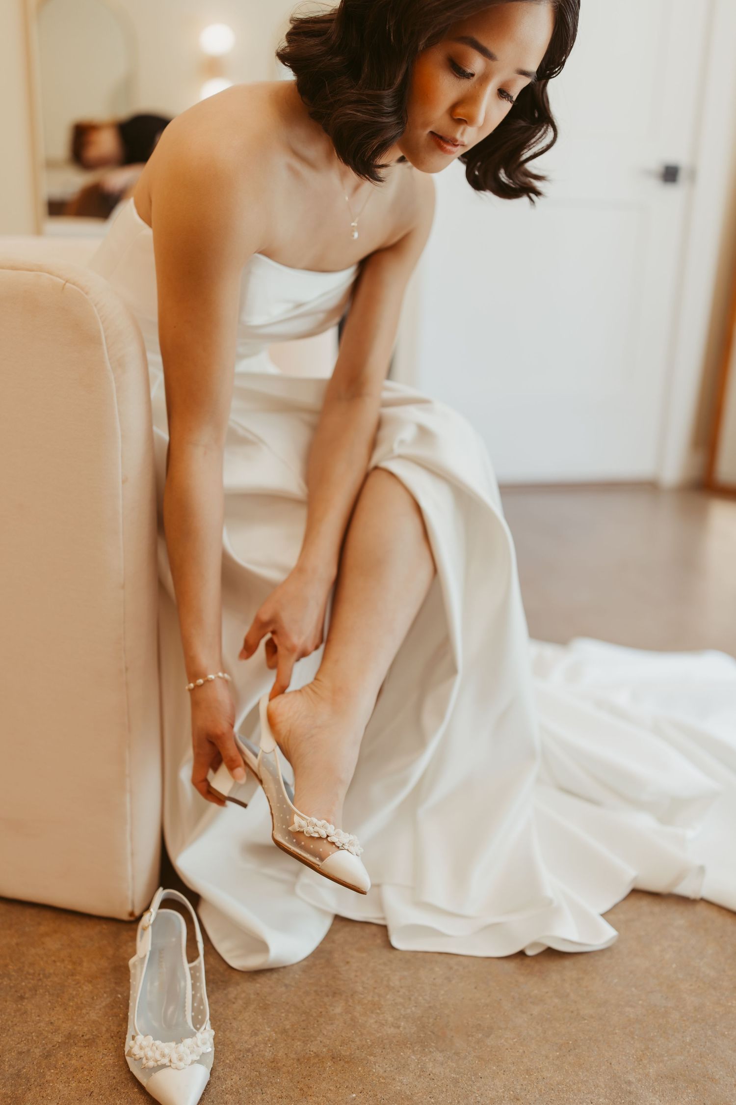 Bride in strapless white satin gown putting on pearl-embellished heels while seated on cream sofa.