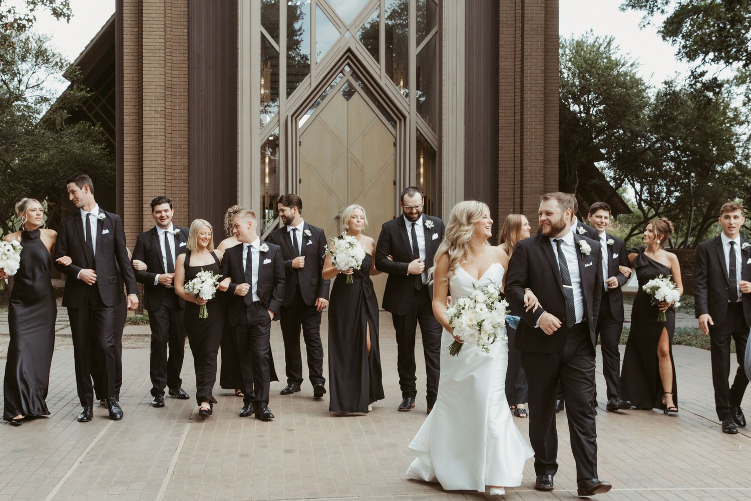 Wedding party in black and white formal attire poses in front of chapel with modern architecture.