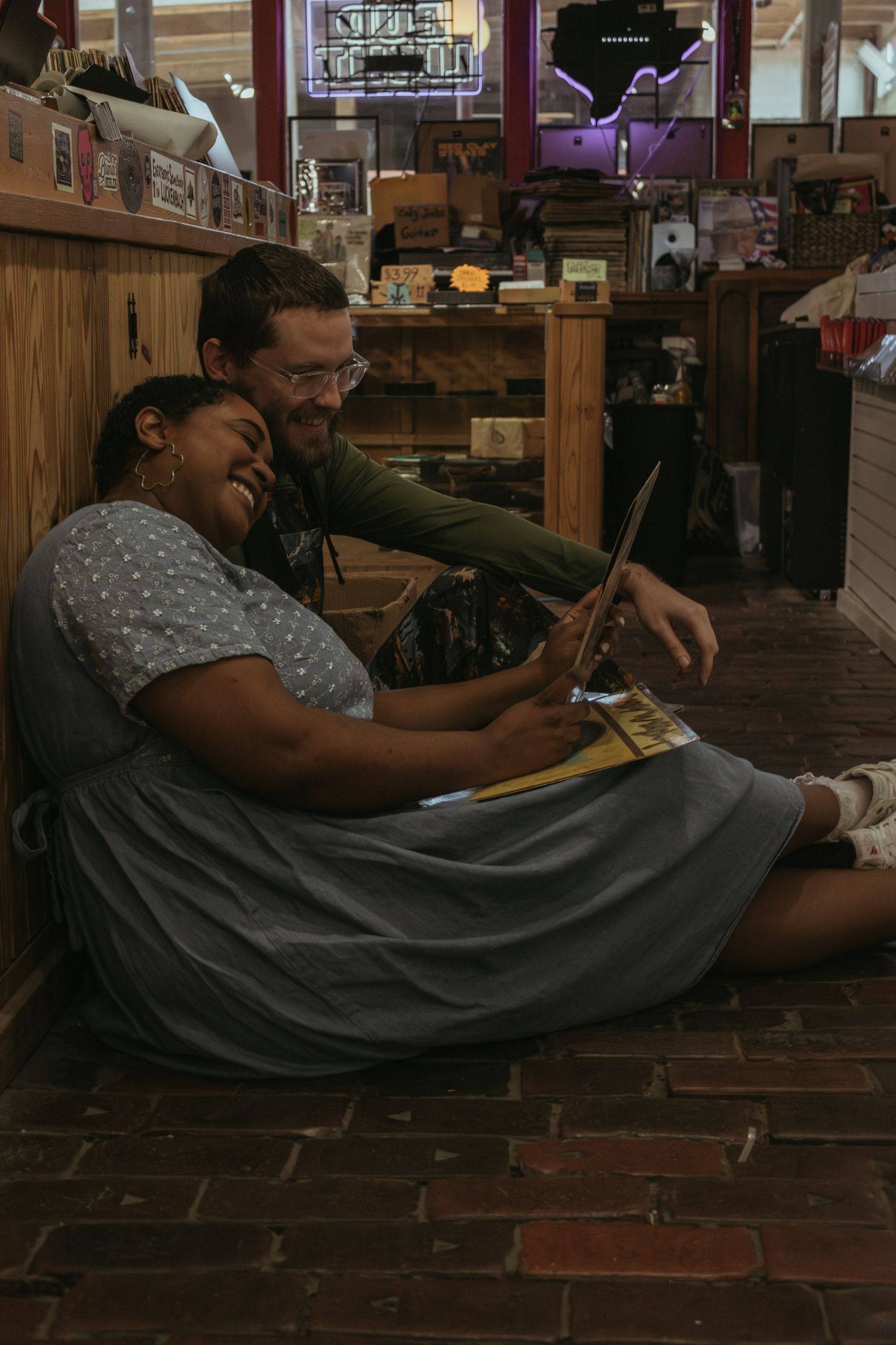 Two people sit on a brick floor together looking at a book in what appears to be a cozy shop interior.