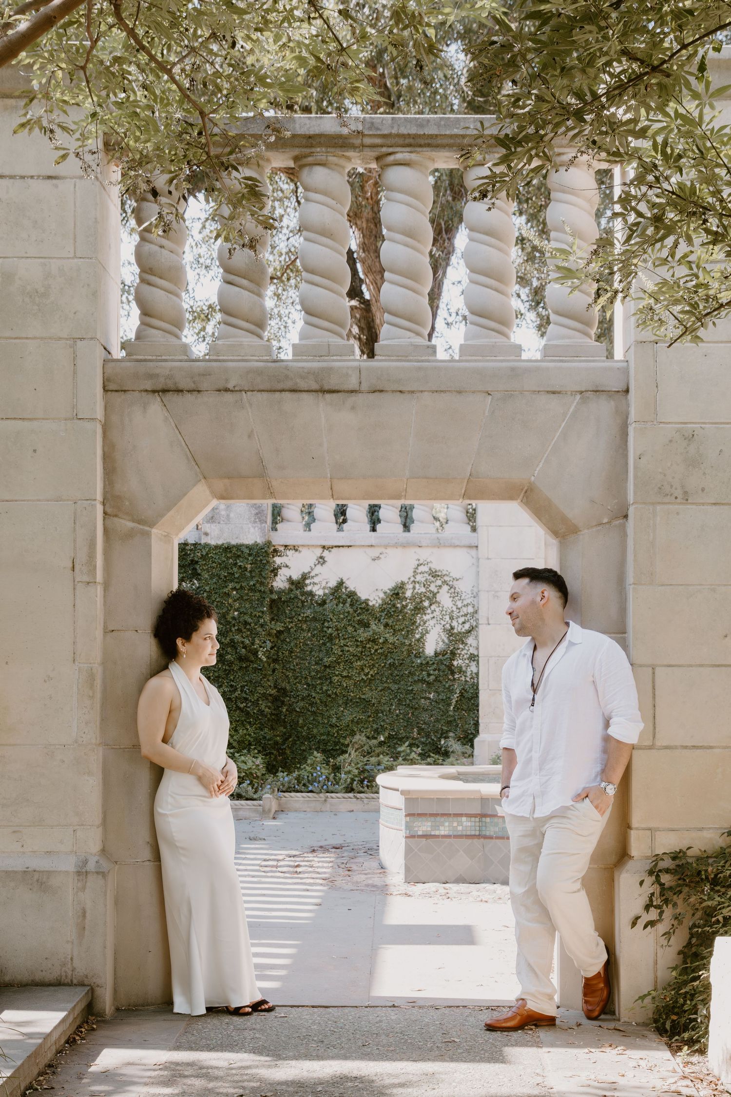 A wedding couple in white outfits sharing intimate moments under a stone archway with hanging lights in an outdoor setting.