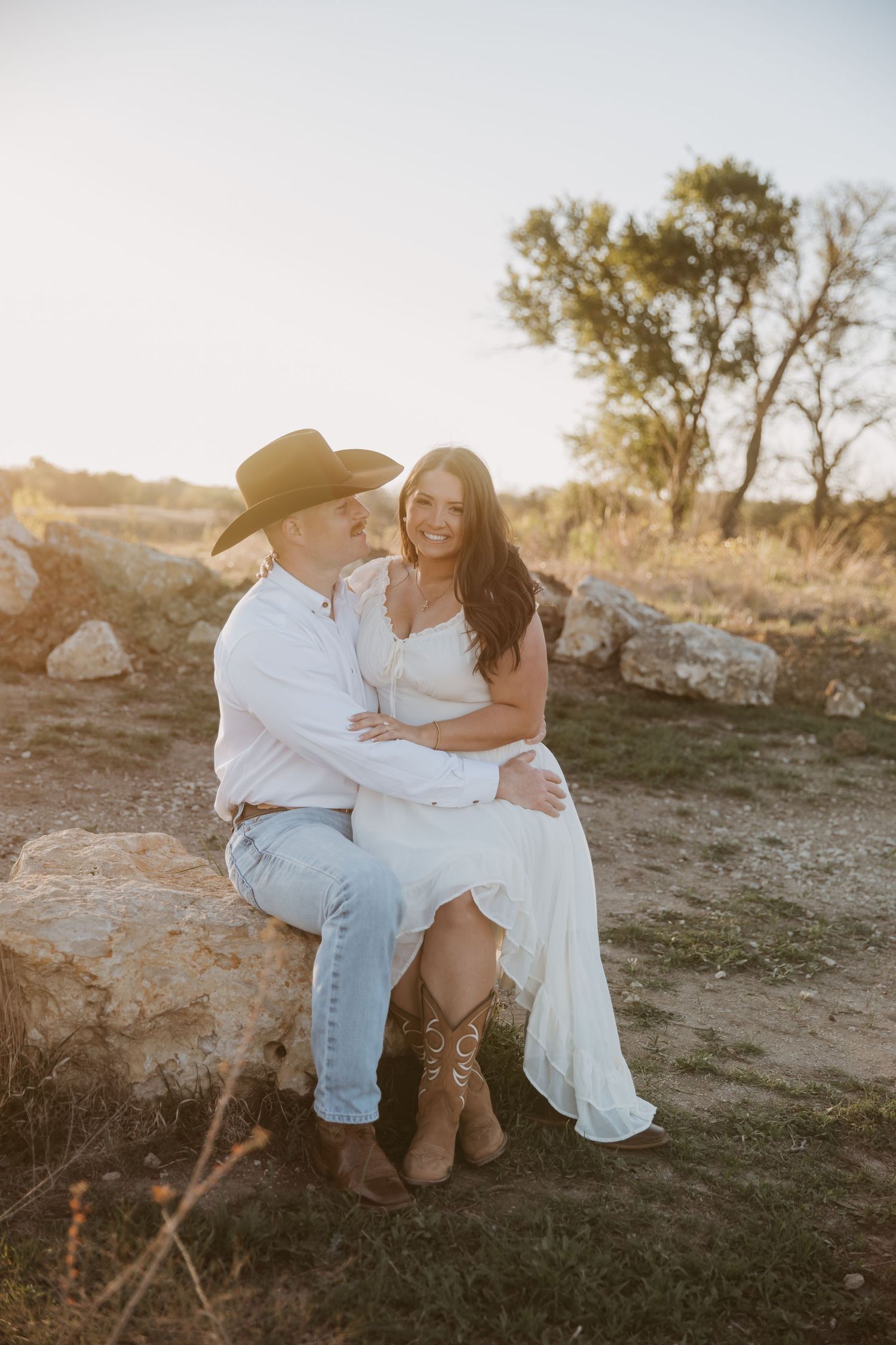 Couple in western attire sitting on rocks at golden hour, woman in white dress and boots, man in cowboy hat and jeans.