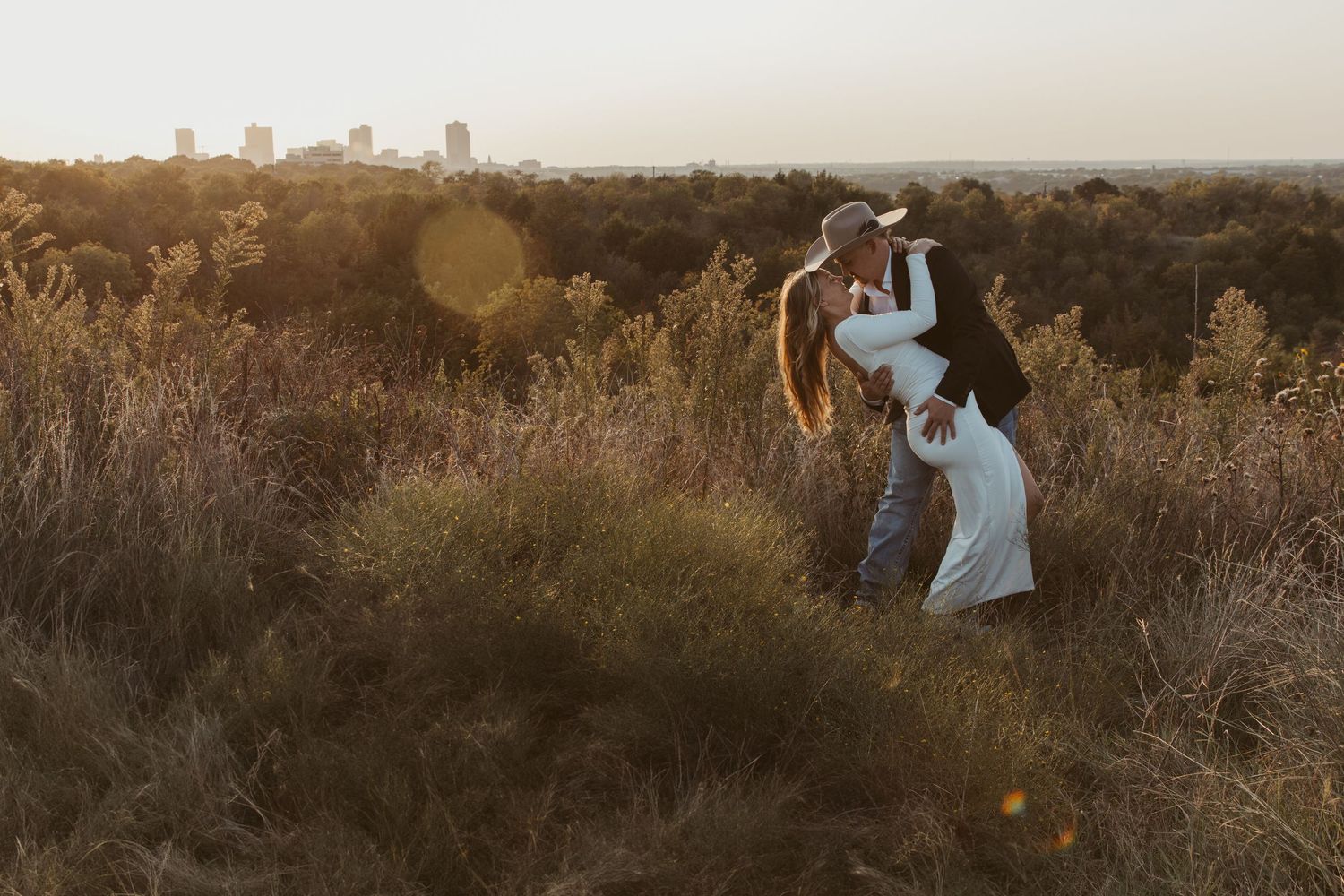 A romantic couple embraces in a golden sunset field surrounded by tall grass and warm autumn light. Engagement photographer DFW