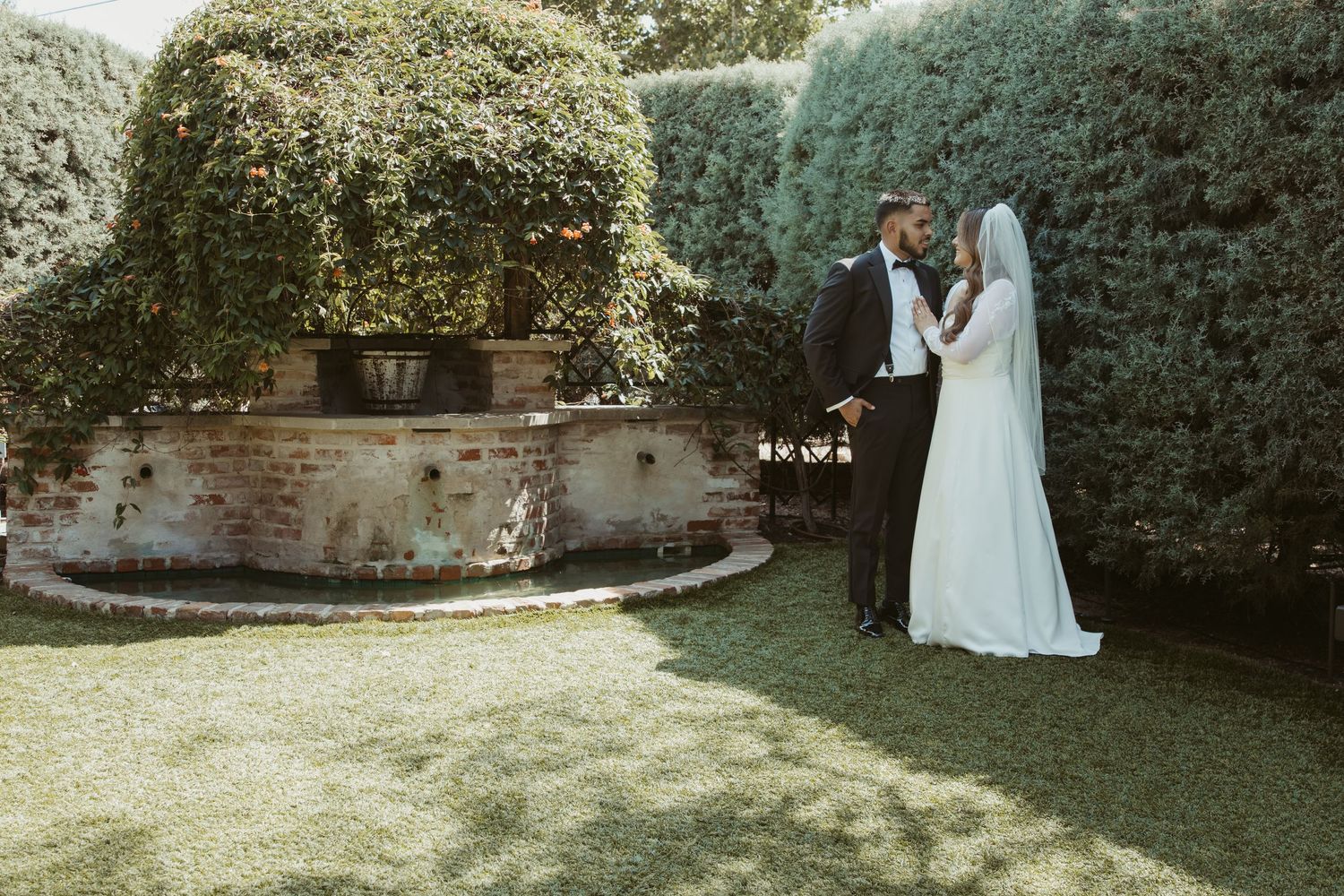 A couple in wedding attire walks through a lush garden pathway with green hedges and trees. Wedding Photographer DFW
